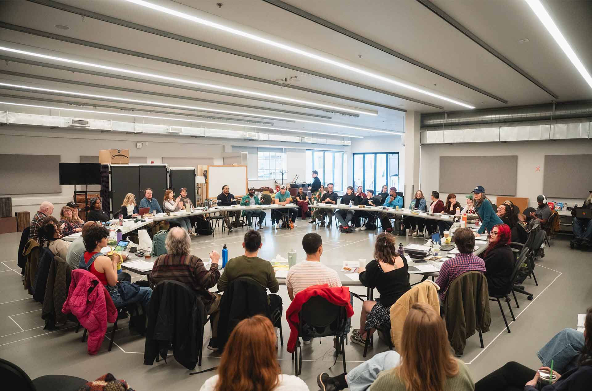 Wide shot of a rehearsal room filled with dozens of people seated in a large circle, with scripts, drinks, and laptops on tables.