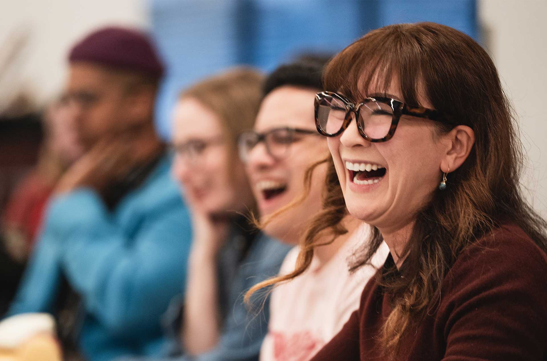 A person in large glasses laughs during a rehearsal, with others beside them also smiling and enjoying the moment.