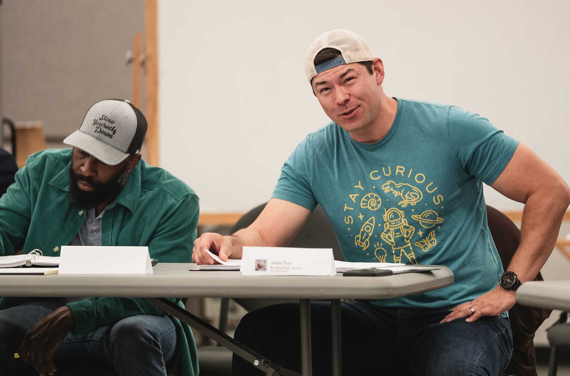 Two people sit at a table reading scripts, one in a “Stay Curious” graphic tee and the other wearing a “Slow Yourself Down” hat.