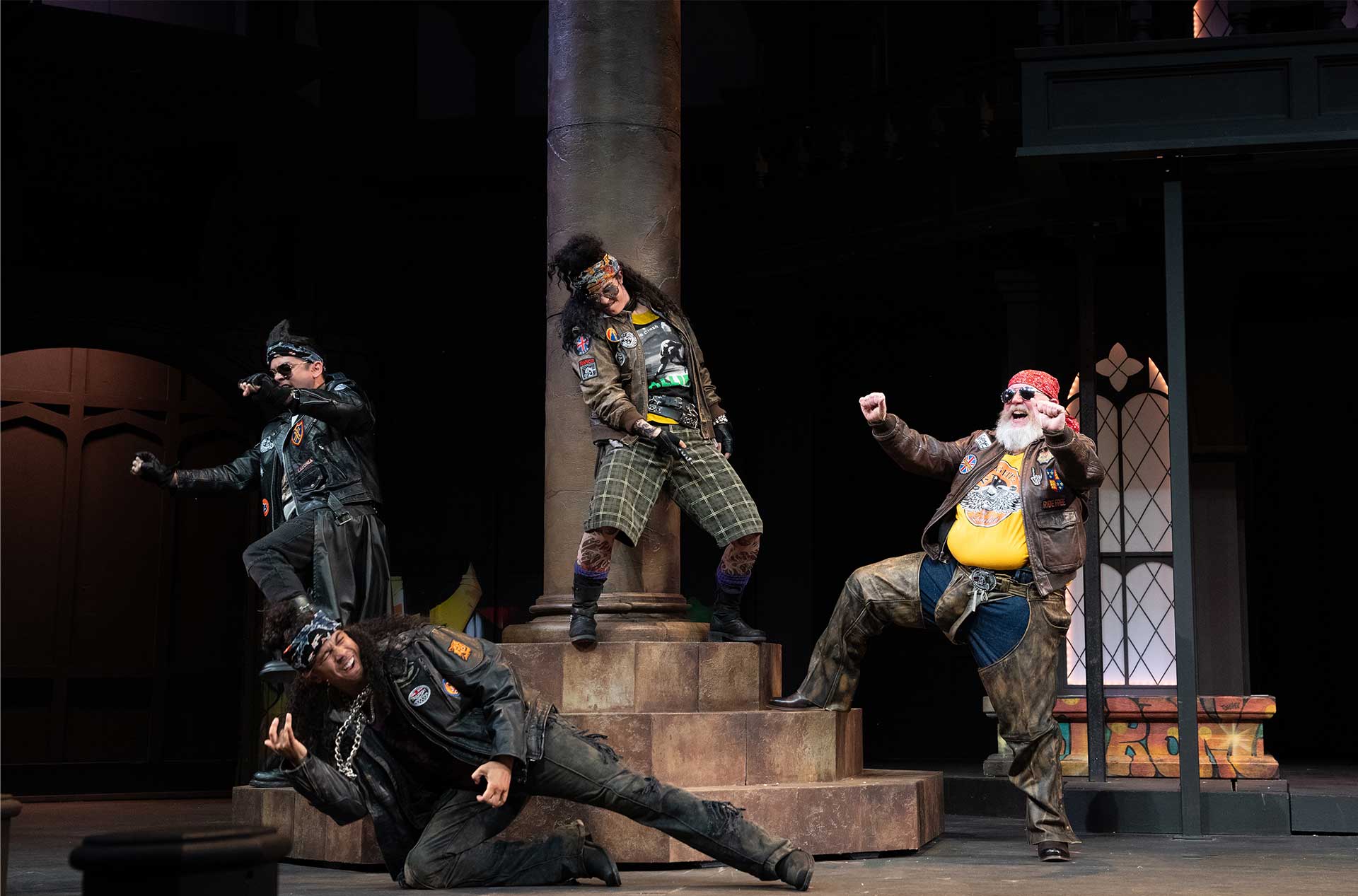 Four performers dressed in rugged biker-style outfits strike exaggerated, energetic poses around a stone column on stage, suggesting a comedic or rebellious group dynamic.