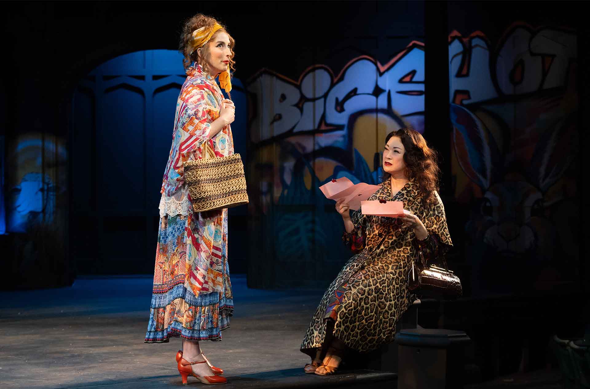One performer stands in a vibrant patchwork dress and headscarf holding a woven bag, while another, seated in leopard print, reads a pink letter with a surprised expression. A mural behind them features graffiti-style lettering.