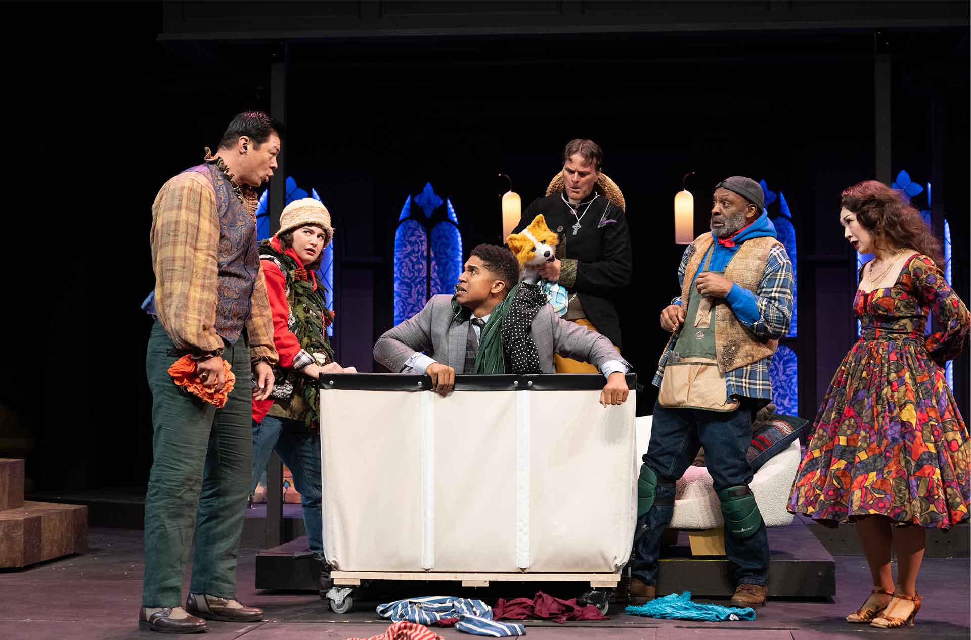 Seven cast members surround a large laundry bin on wheels, with one person inside the bin looking up in alarm. Another character holds a fox puppet, and various costumes suggest playful or chaotic storytelling.