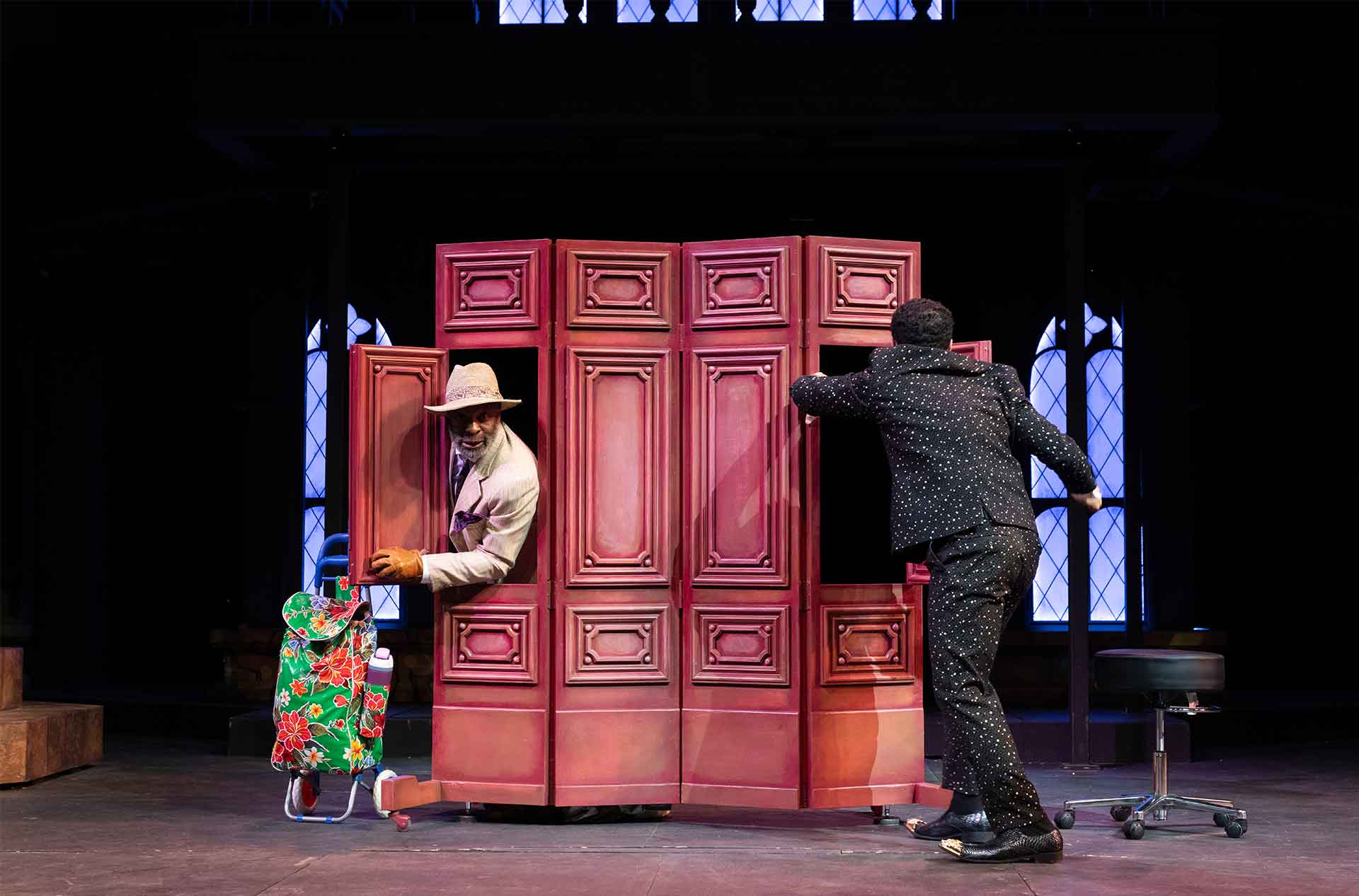 One performer peers out from behind a large, red folding screen holding a floral shopping cart, while another in a dotted suit pushes the panels, suggesting a humorous or mistaken-identity moment.
