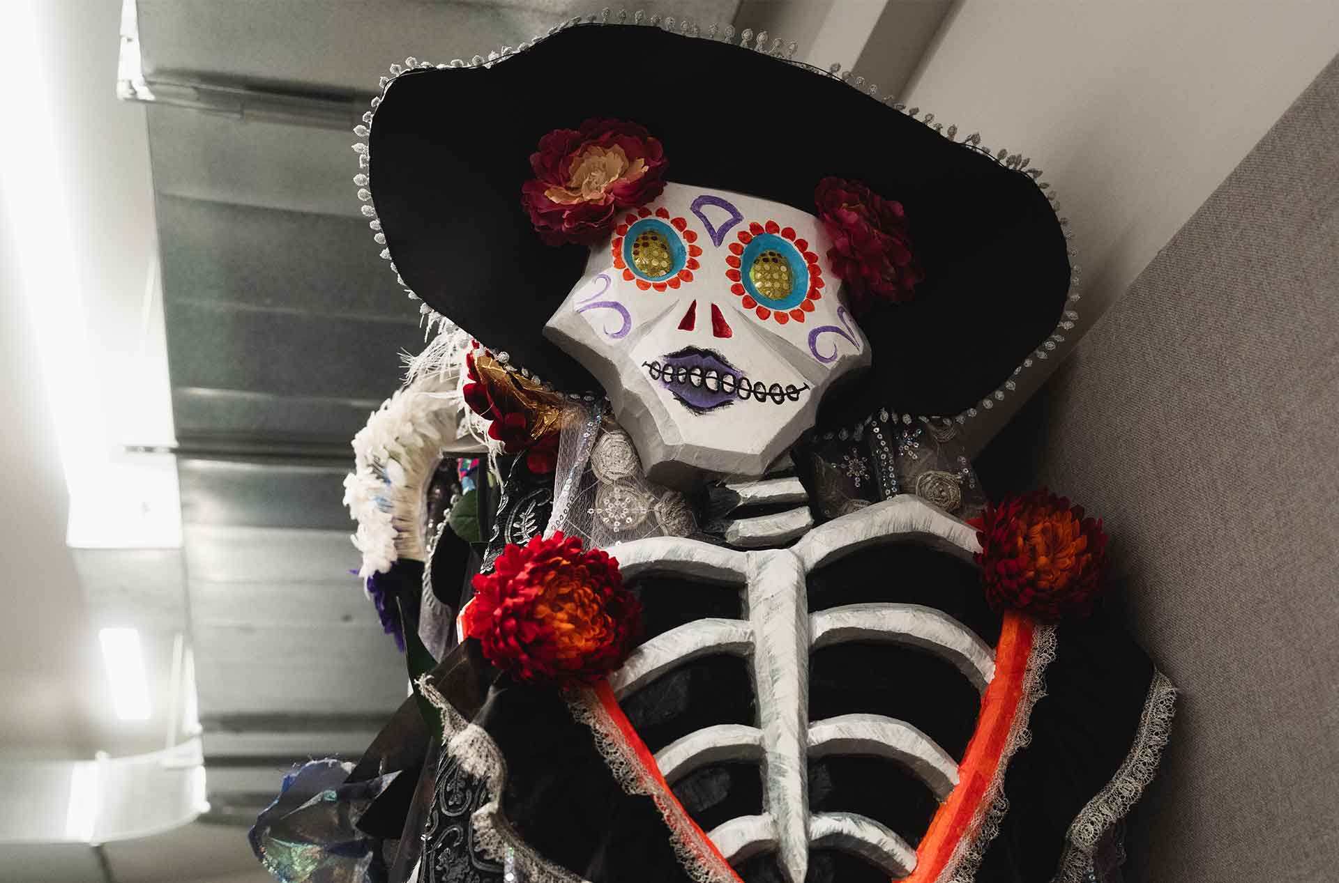 Decorative Día de los Muertos-style skeleton figure with a black hat, colorful face paint, and flower adornments, displayed in the rehearsal space.