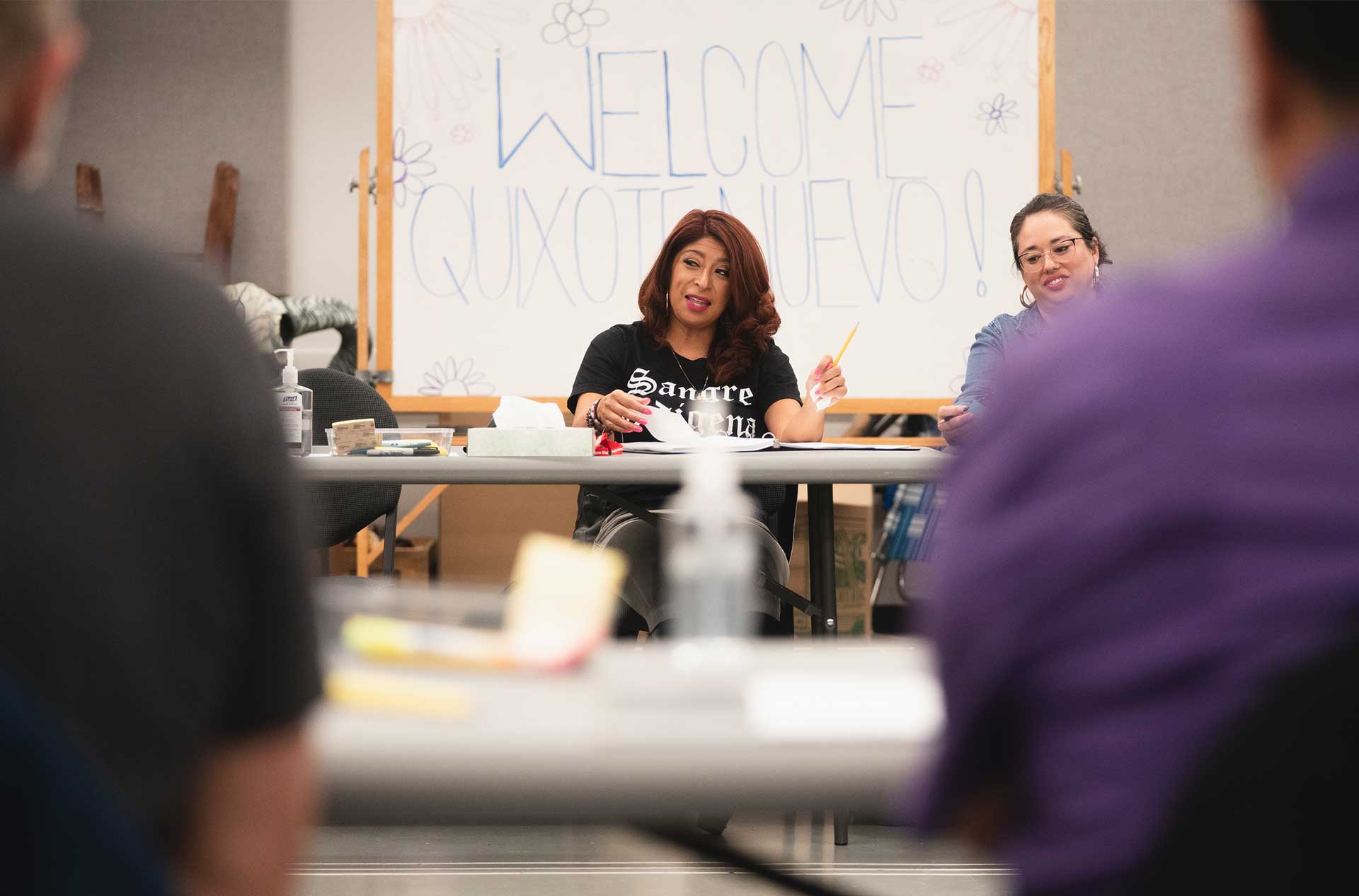 Two people seated at a rehearsal table, one speaking with expressive gestures, in front of a sign reading “Welcome Quixote Nuevo!” decorated with hand-drawn flowers.