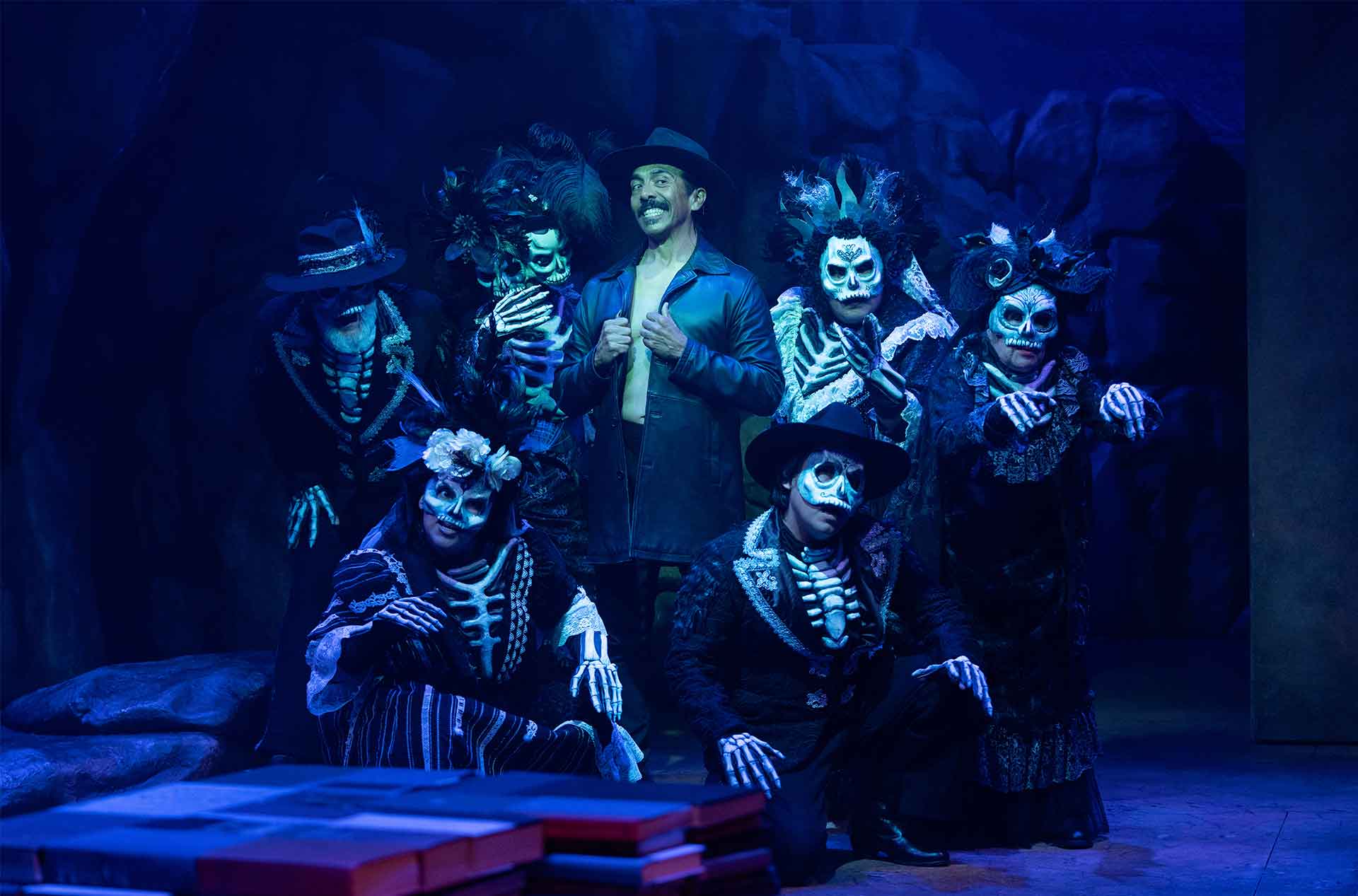 Group of actors in skeleton makeup and dark costumes pose around a central character in a black leather coat, under blue stage lighting.