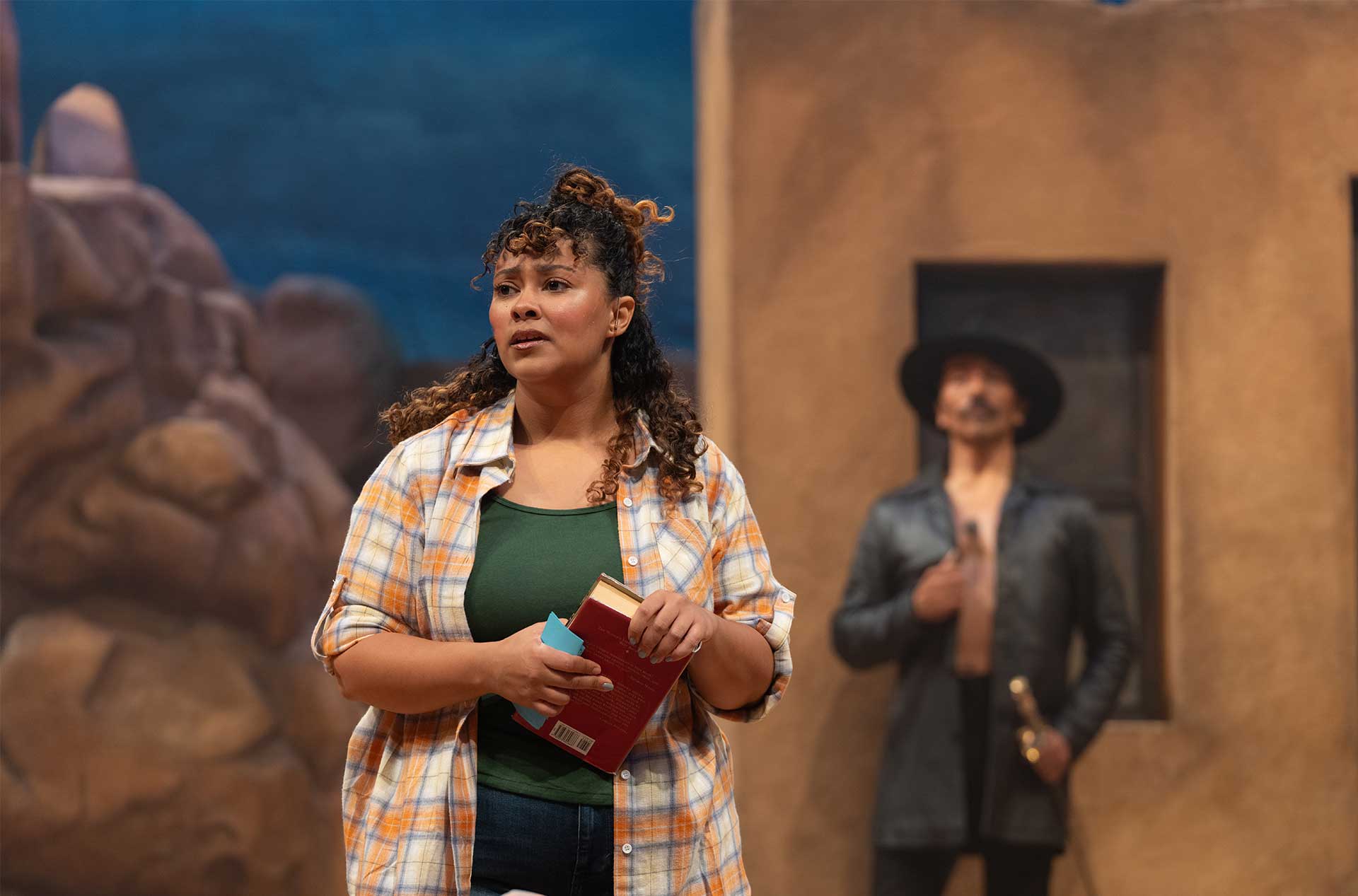 Actor in a plaid shirt holding books, standing in front of a painted desert backdrop with another character in a dark outfit and hat blurred in the background.