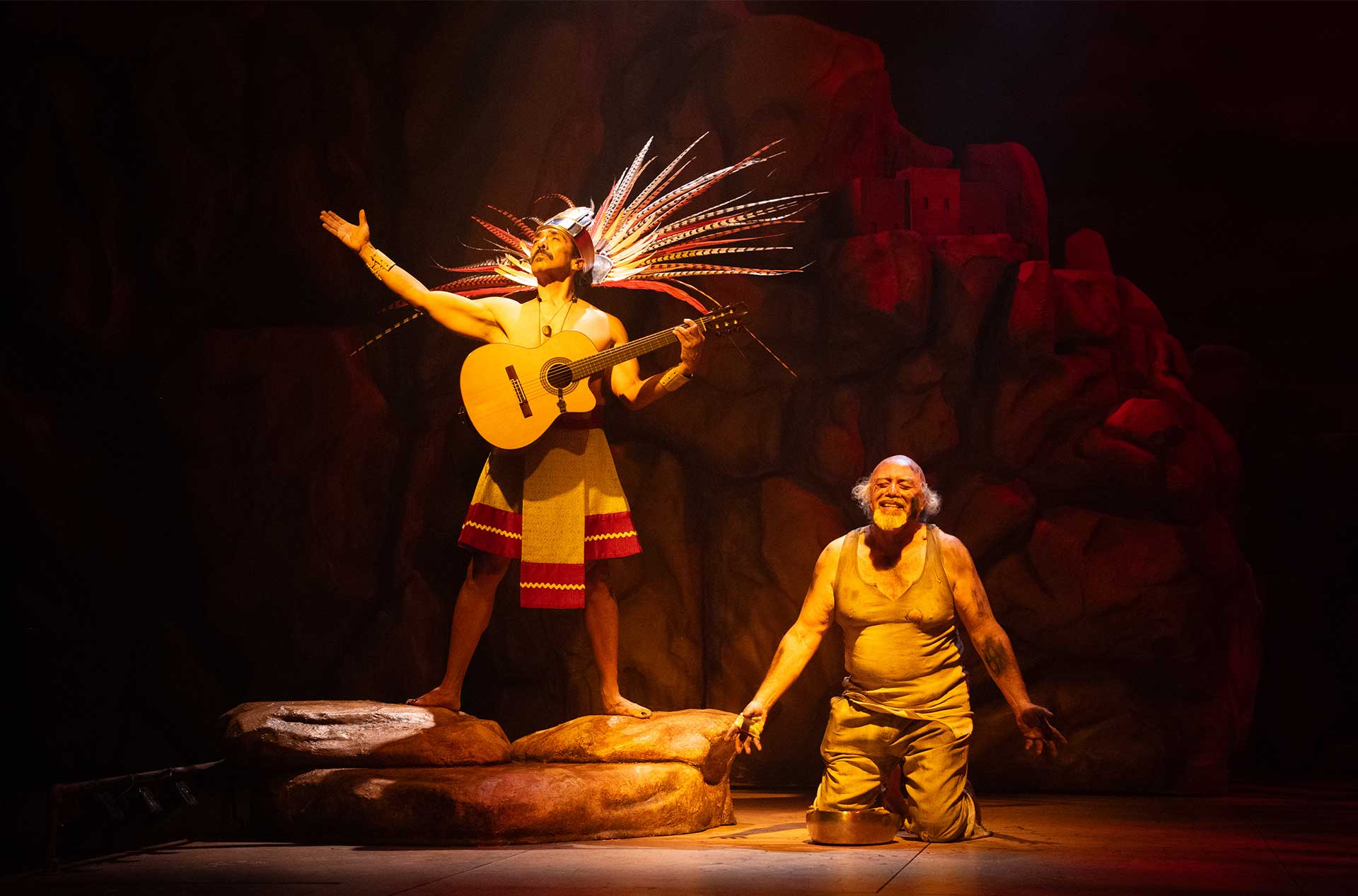 Performer in a feathered headdress plays guitar on a rock formation while another character kneels beside them, both lit by warm stage lights.