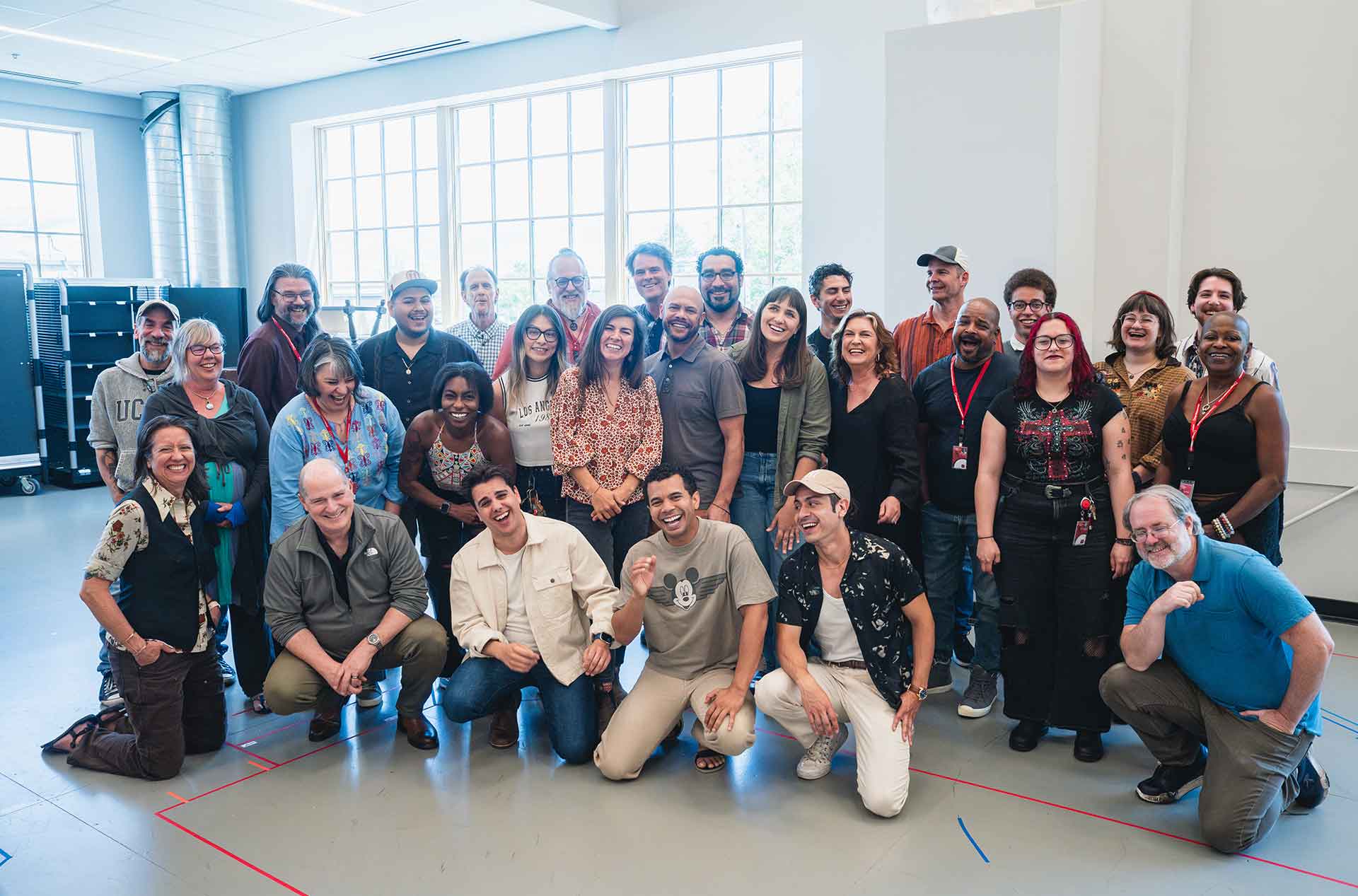 A group of about thirty people stands closely together, smiling and posing for a group photo in a bright indoor space with large windows behind them.