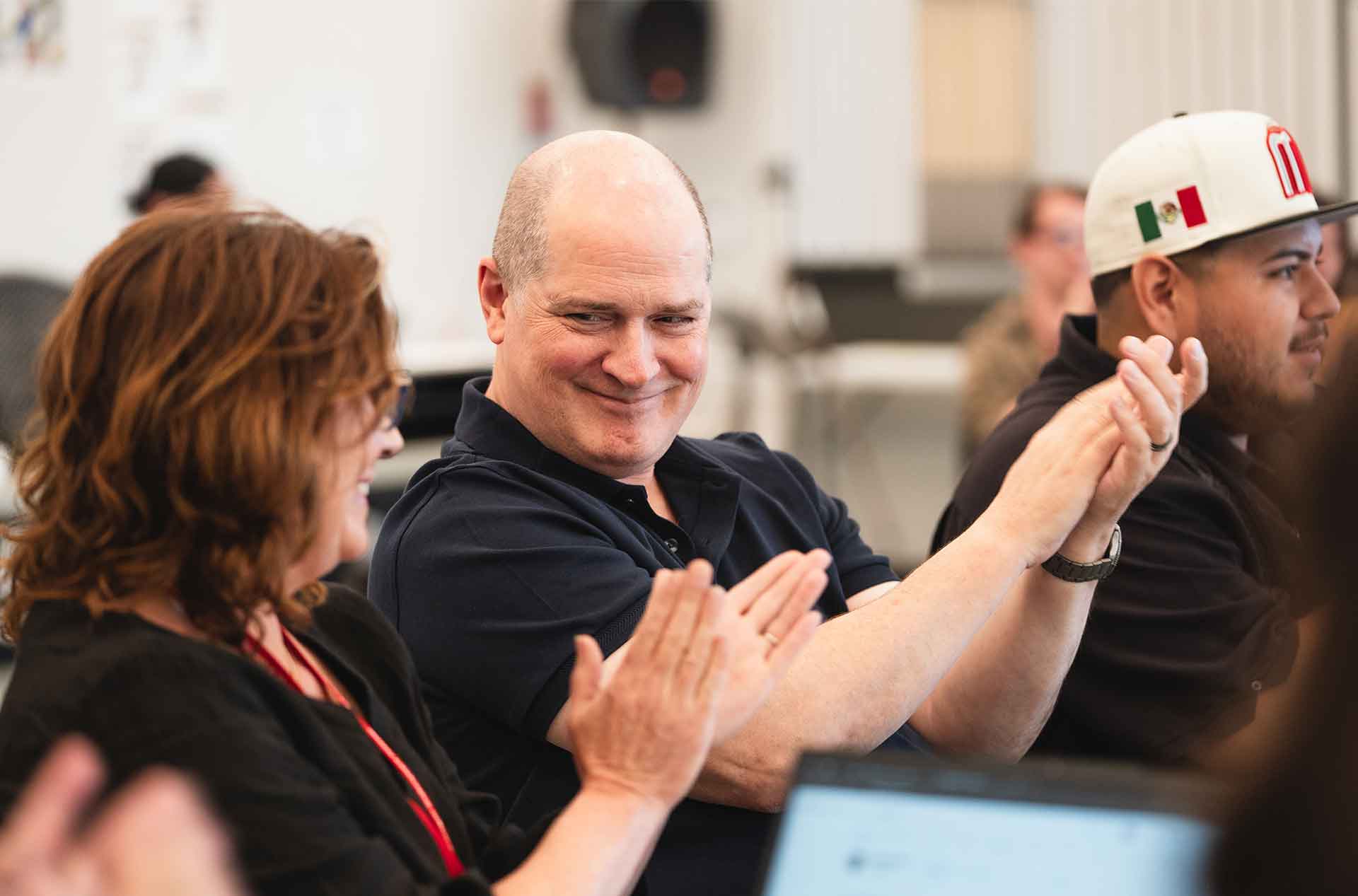 Two people seated next to each other clap while smiling, one glancing sideways with a playful expression; others sit around them in a bright room.