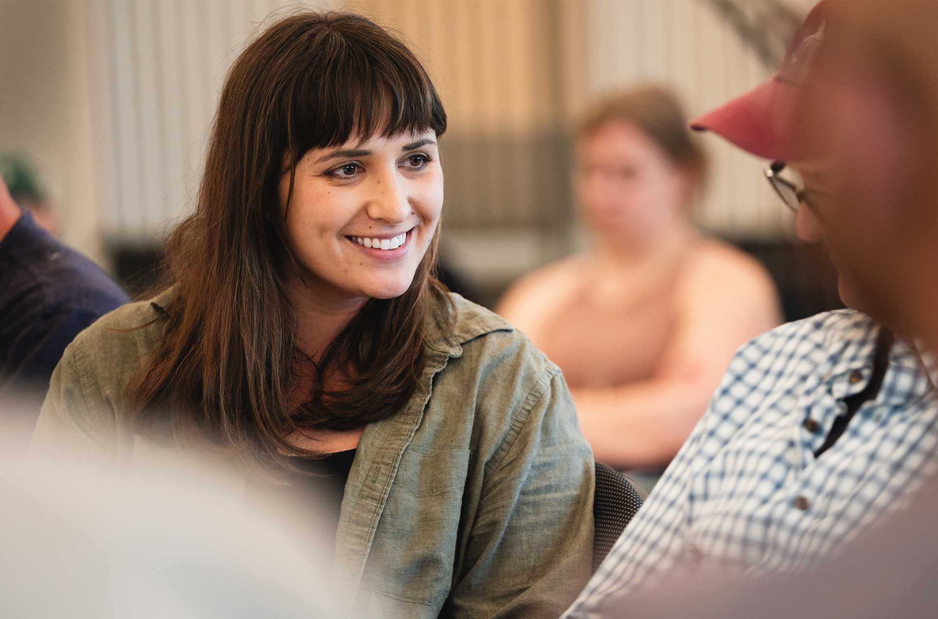 A person with straight dark hair smiles warmly while listening to someone speak in a group setting; blurred people in the background create a soft focus.