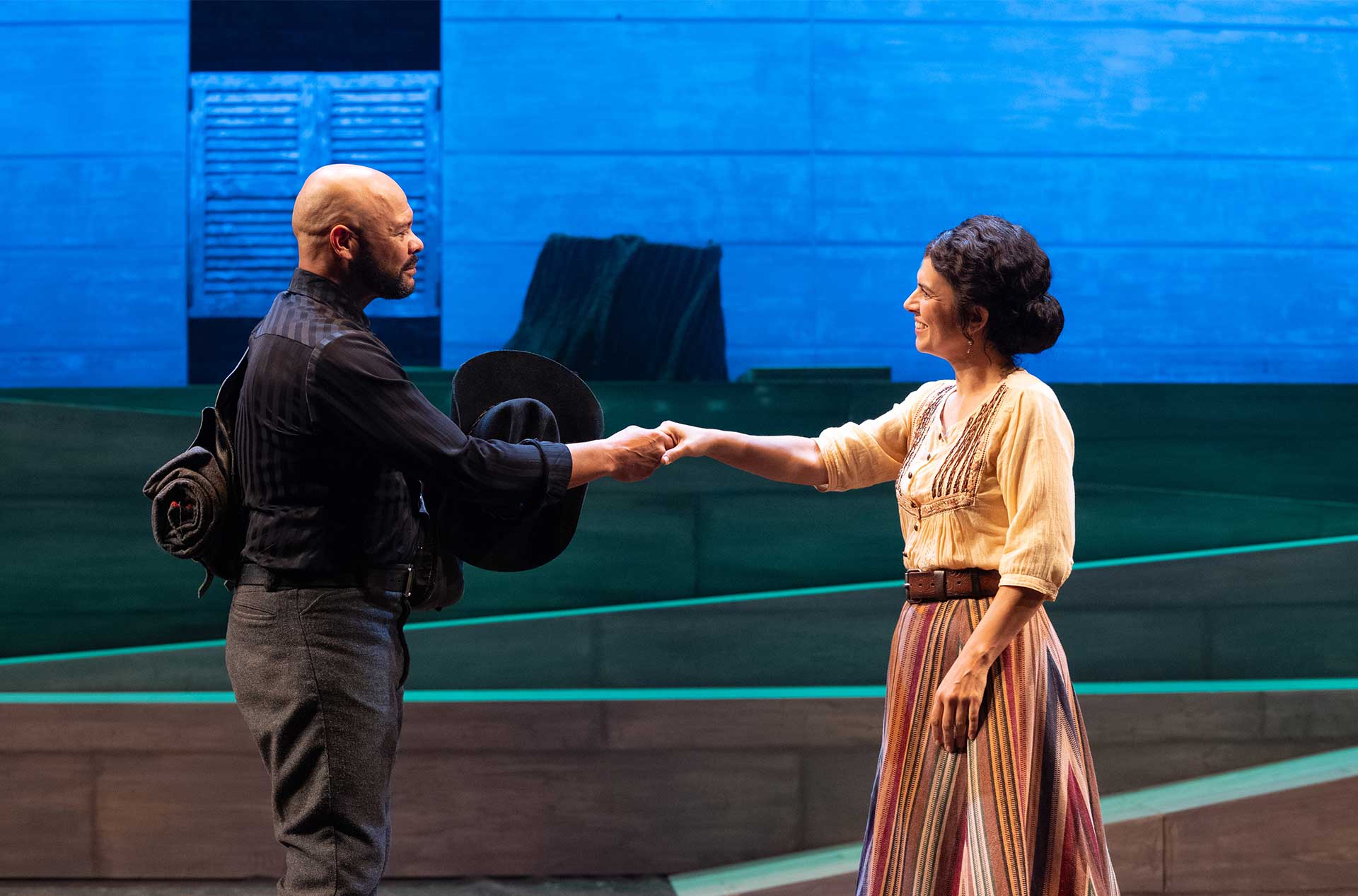 Two performers in period costumes shake hands onstage, with a blue-lit backdrop and wooden set pieces behind them.