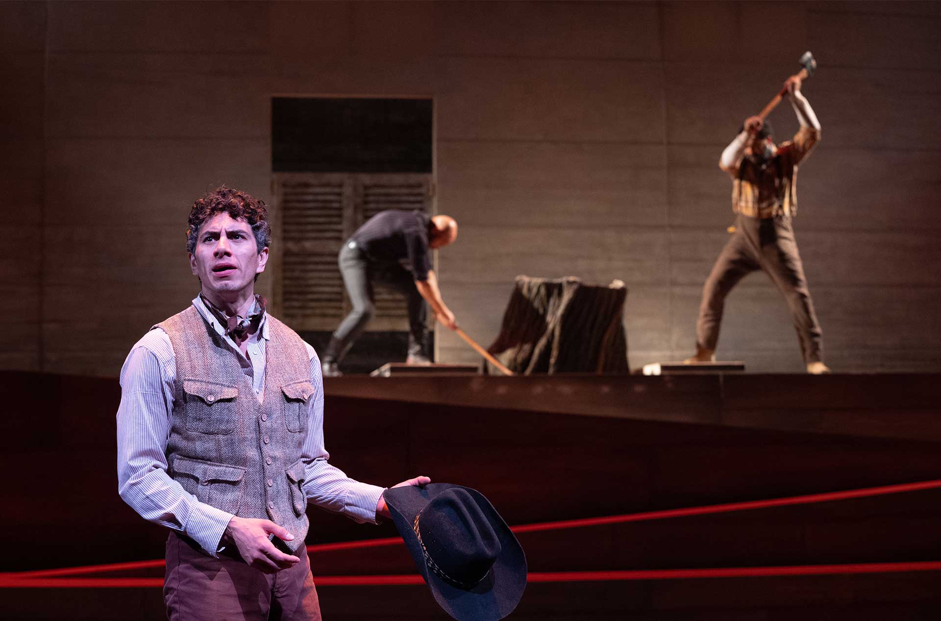 A performer in a vest holds a black hat in the foreground while two others work with an axe and saw in the background.