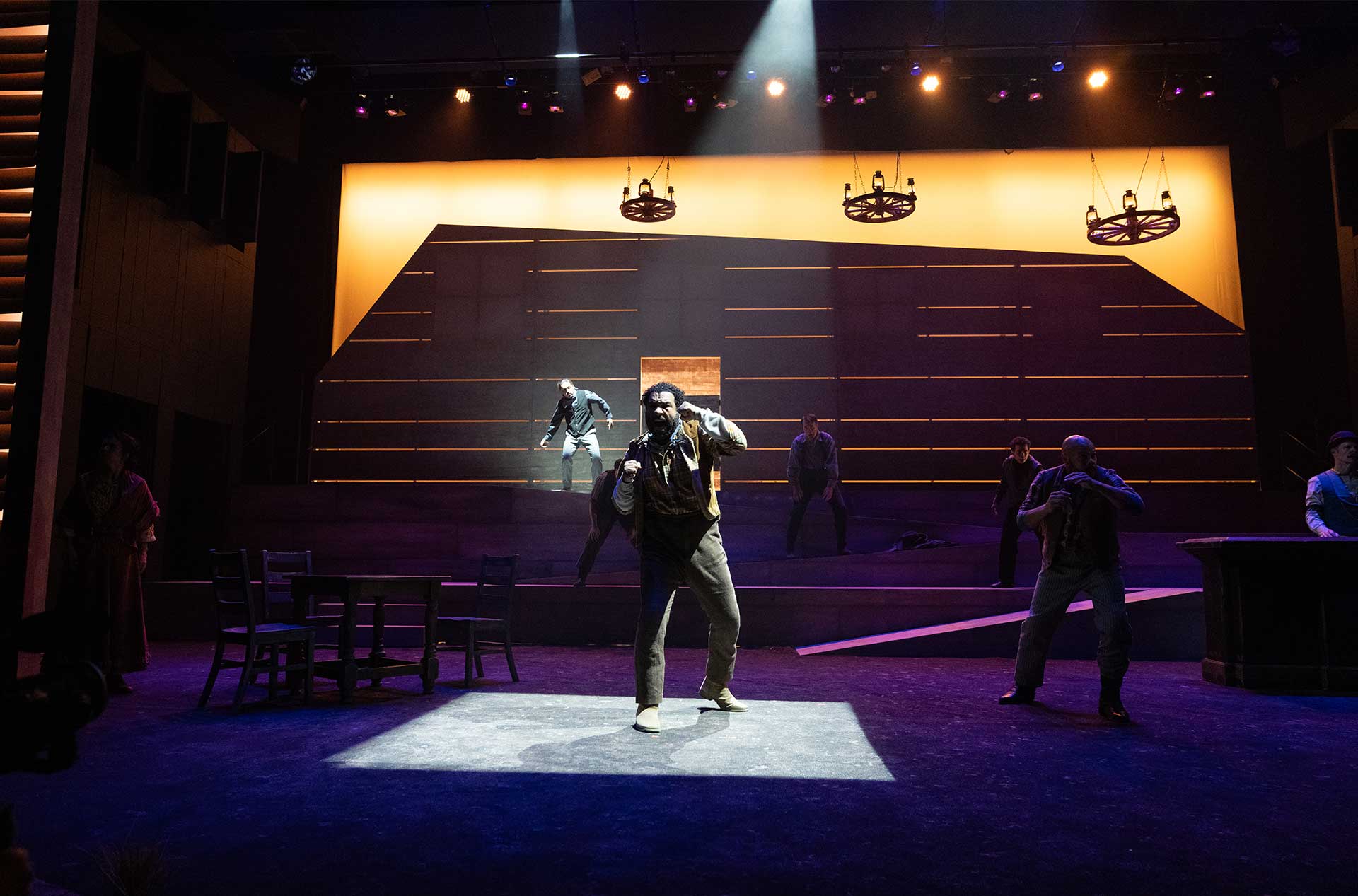 A dramatic spotlight centers a performer gesturing intensely, surrounded by others on a stylized western set with warm backlighting.
