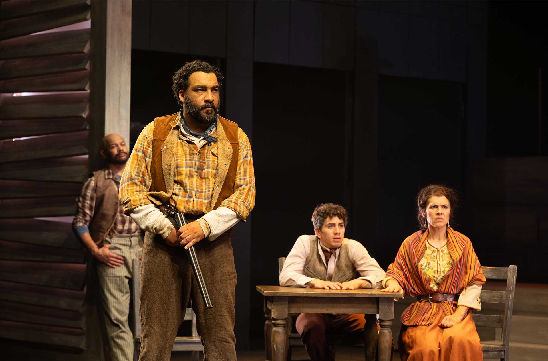 A performer holding a shotgun stands in front of three seated performers who appear tense or concerned.