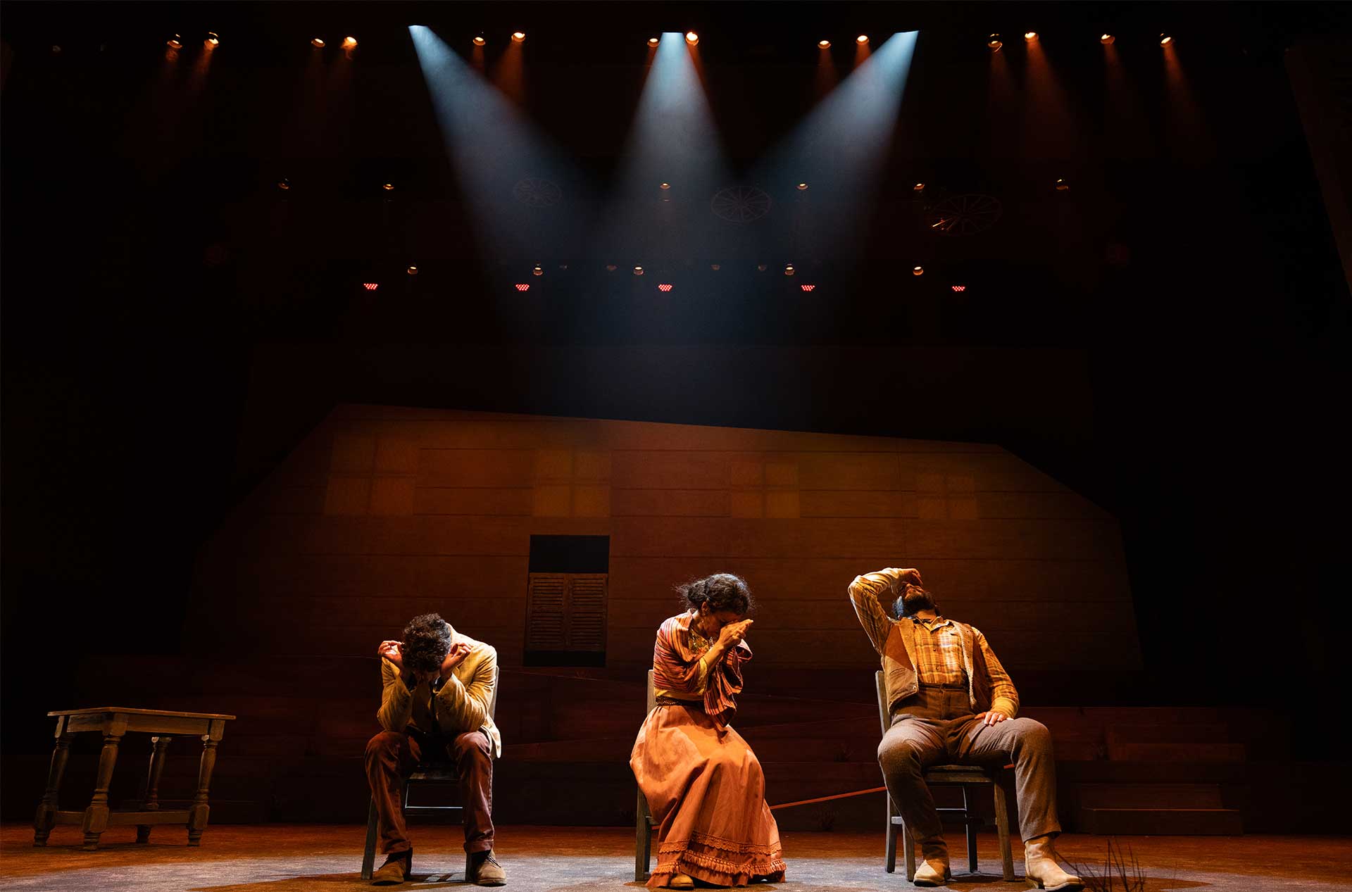 Three performers sit with heads bowed and arms raised, under dramatic lighting and a dark set.