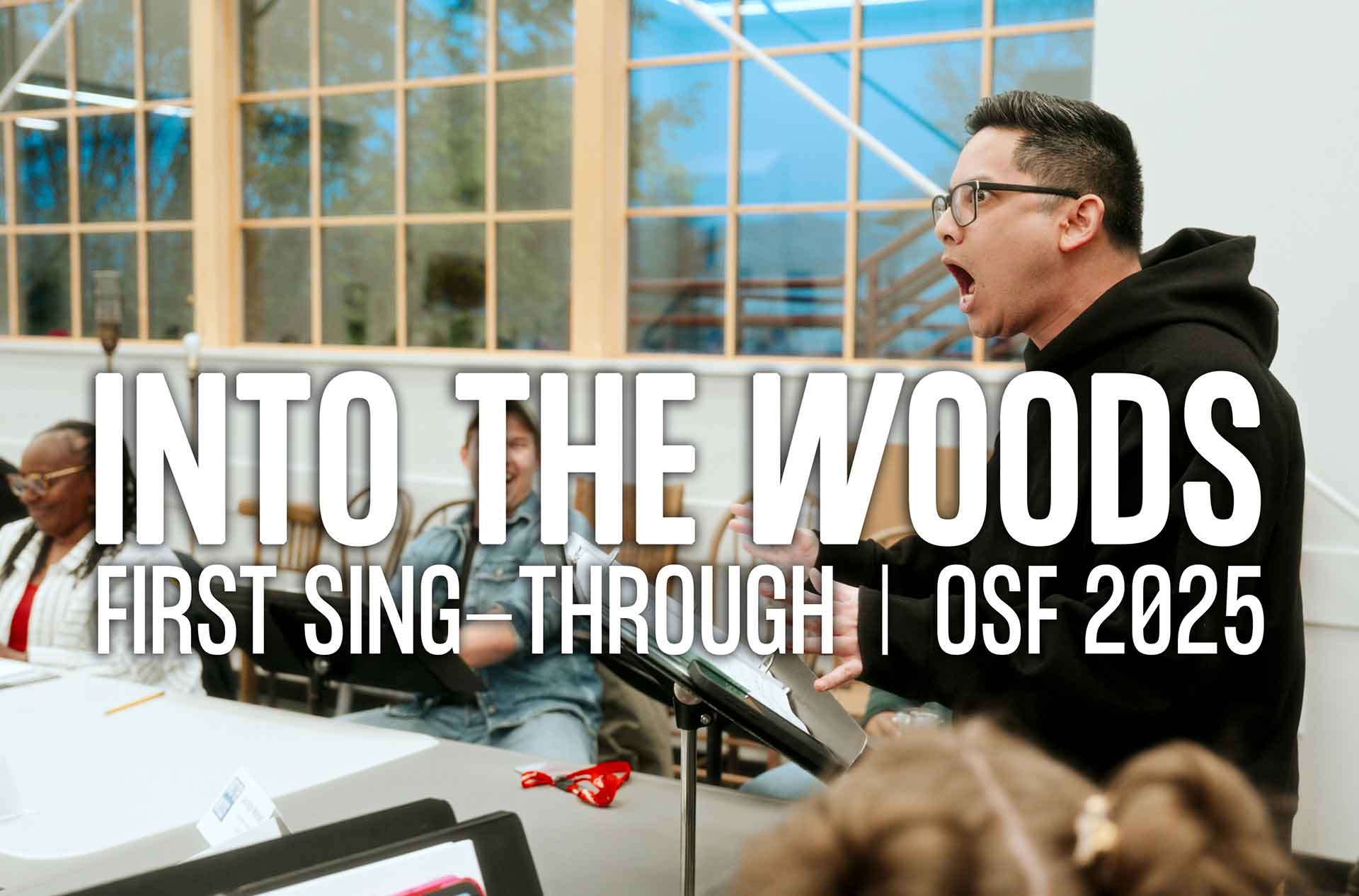 A rehearsal scene for OSF 2025's Into the Woods, showing a passionate man in a black hoodie singing with intensity, while others sit at a table with scripts, smiling and engaged. Large text reads: “INTO THE WOODS – FIRST SING-THROUGH | OSF 2025.