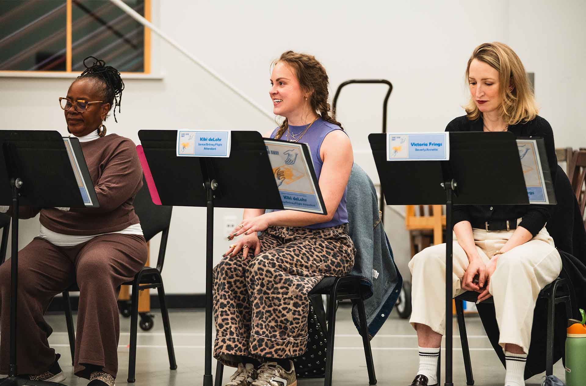 Three participants sit with music stands, listening and smiling during rehearsal. Name cards are clipped to the stands, indicating an organized group session.