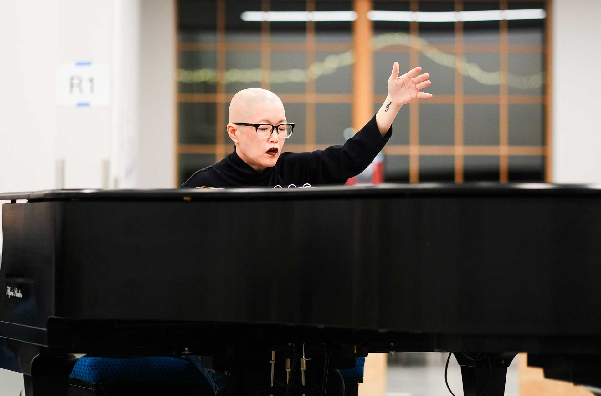 A Music Director sits at a piano with one hand raised mid cue. The gesture suggests active direction during a musical rehearsal.