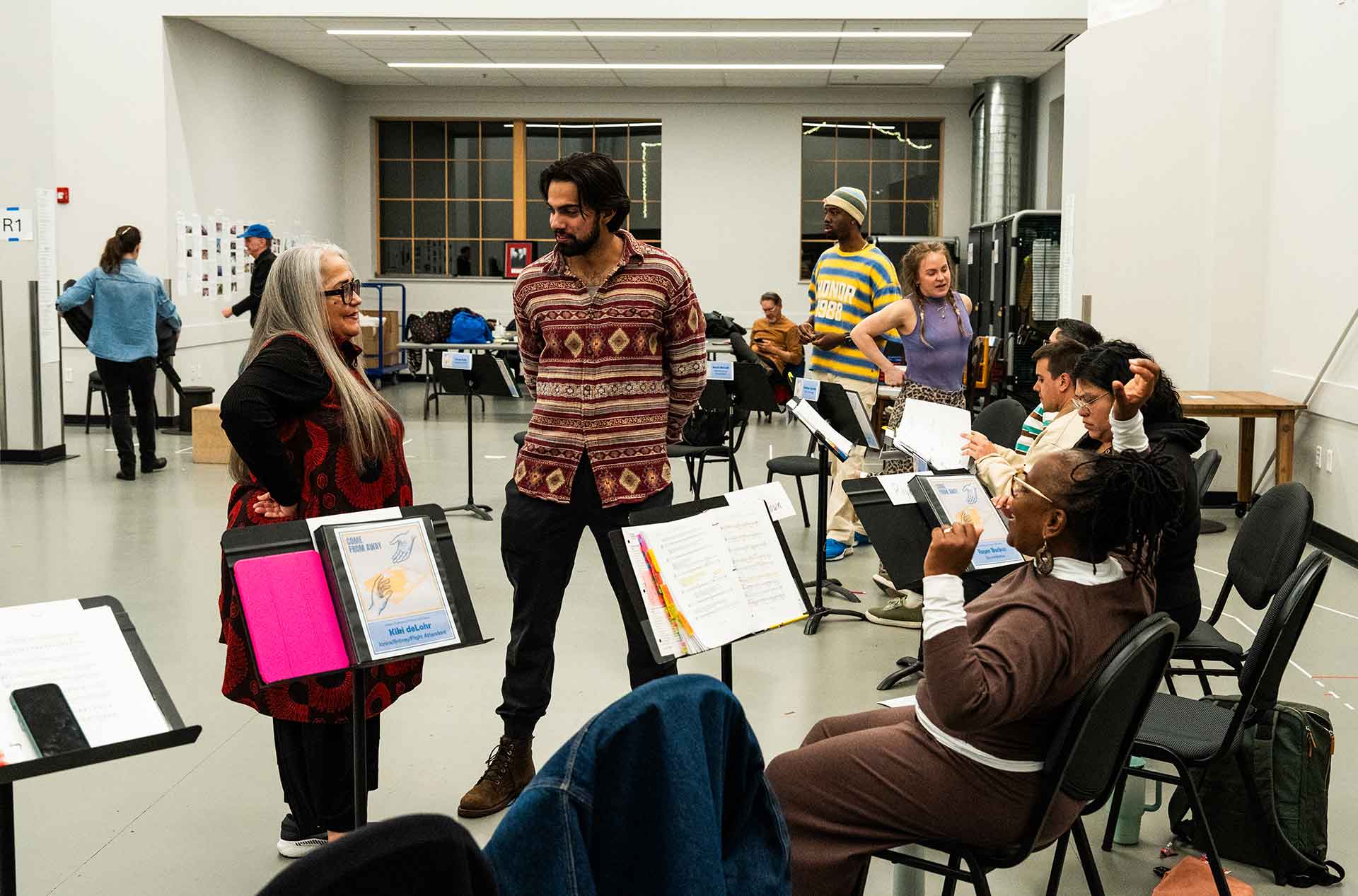 Two people stand talking among seated participants during a break in rehearsal. Music stands, chairs, and others preparing materials fill the room behind them.