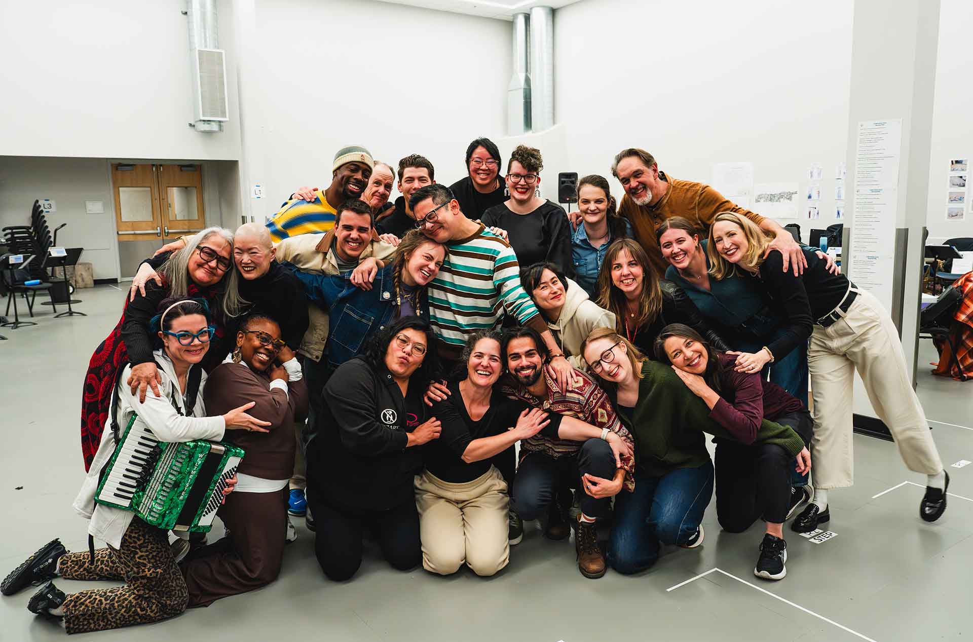 A large group poses closely together in a rehearsal space, smiling and leaning into one another. The image conveys a sense of community and shared accomplishment after a group activity.