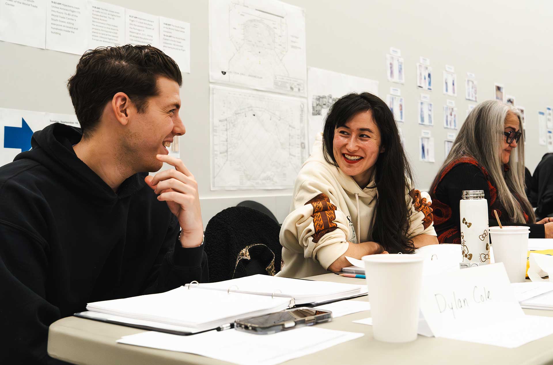 Two participants sit at a table smiling and talking during a working session. Papers, binders, and cups are spread across the table, suggesting collaborative discussion.