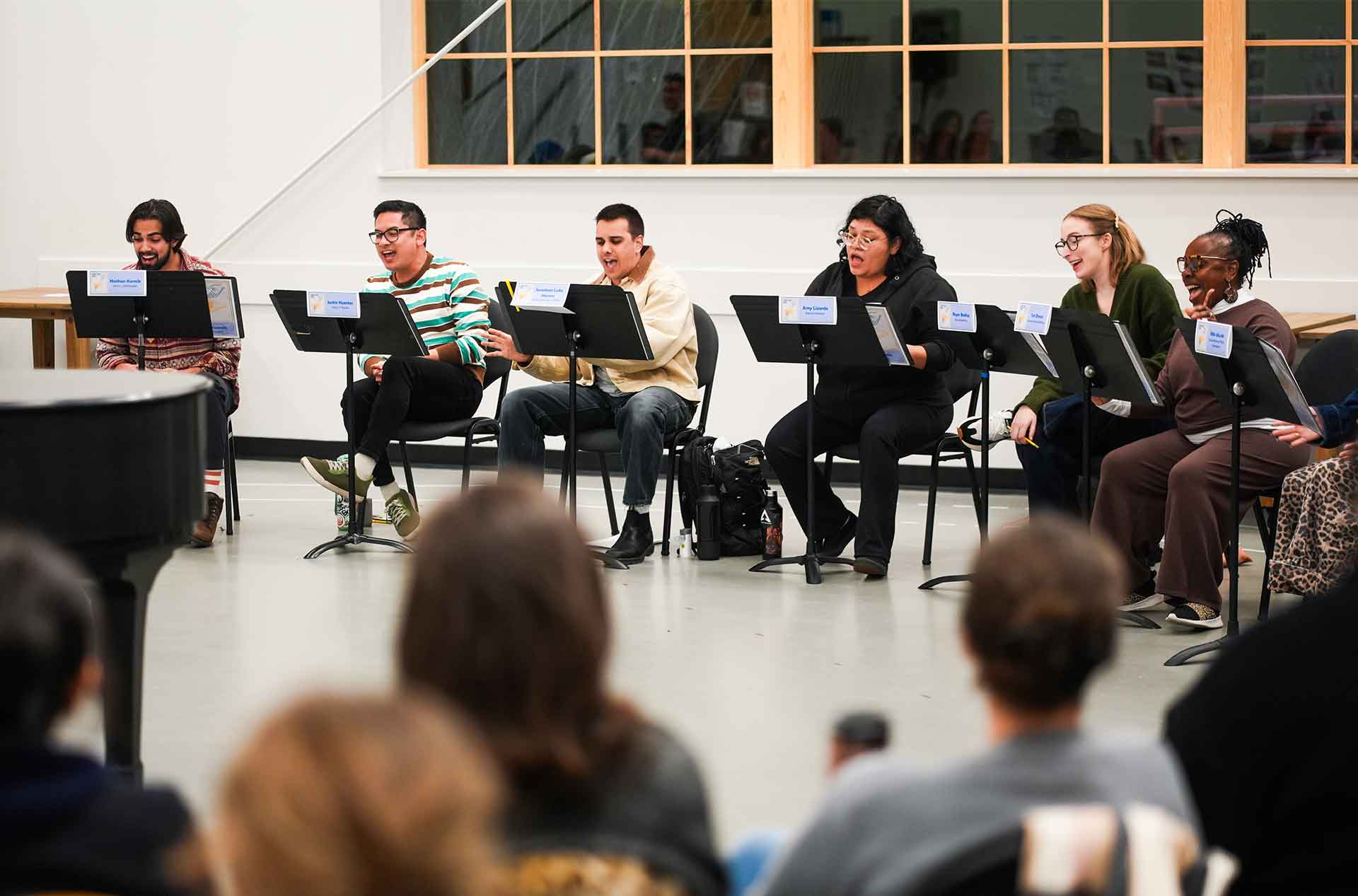 A row of participants sit on chairs singing from music stands during a rehearsal. An audience is visible in the foreground, watching attentively.