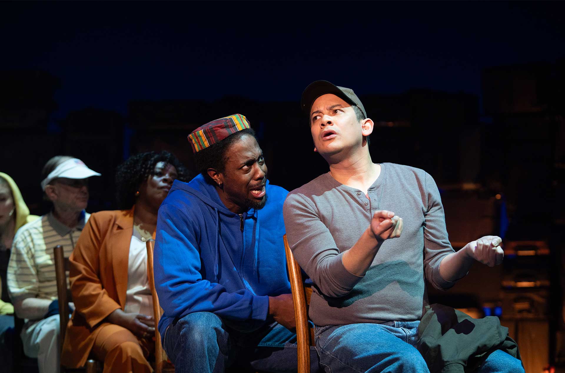 Two people sit close together in a row of chairs, one gesturing animatedly while others watch from behind.