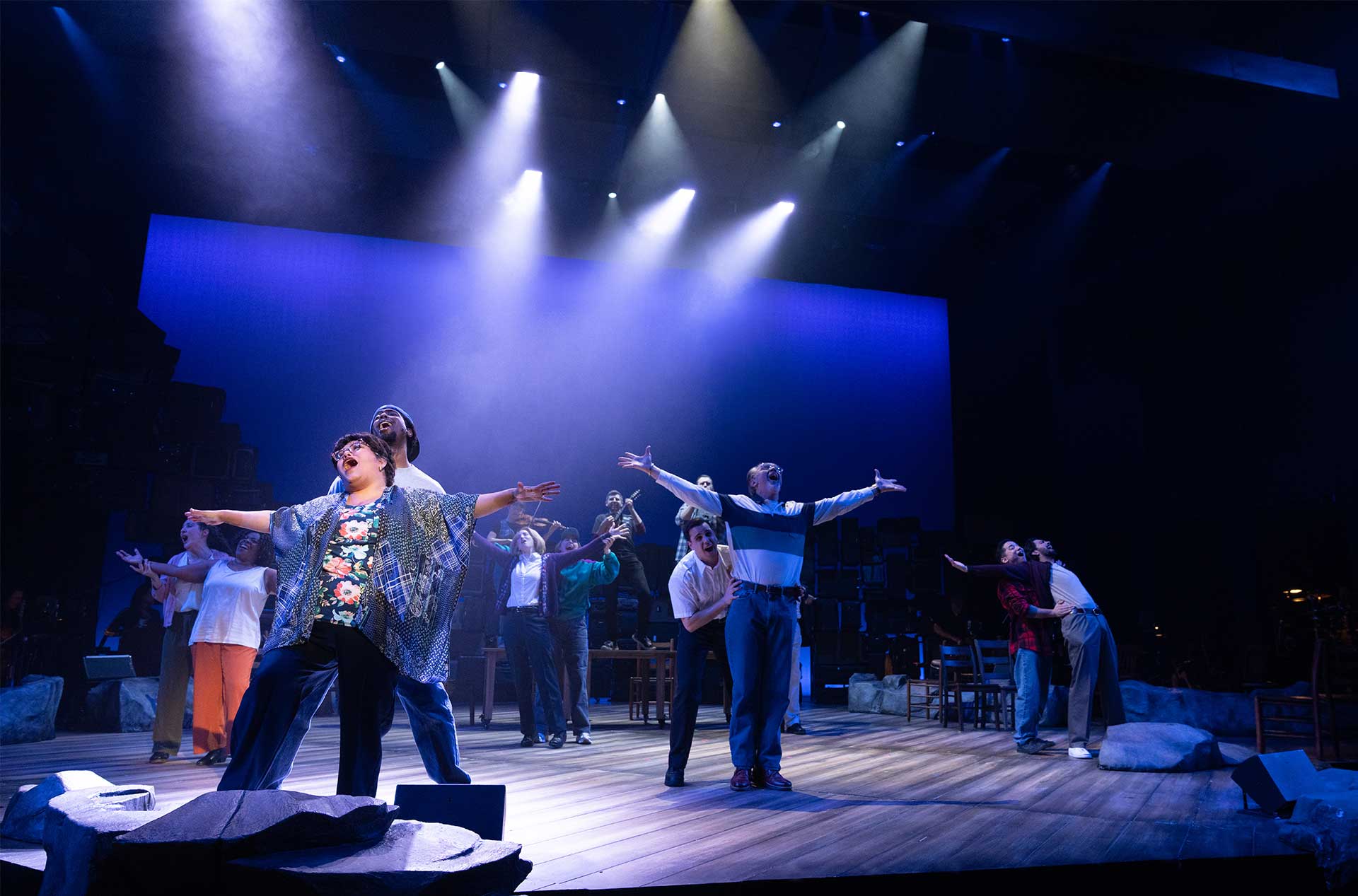 A large ensemble performs with arms outstretched under bright stage lights in a wide theatrical scene.