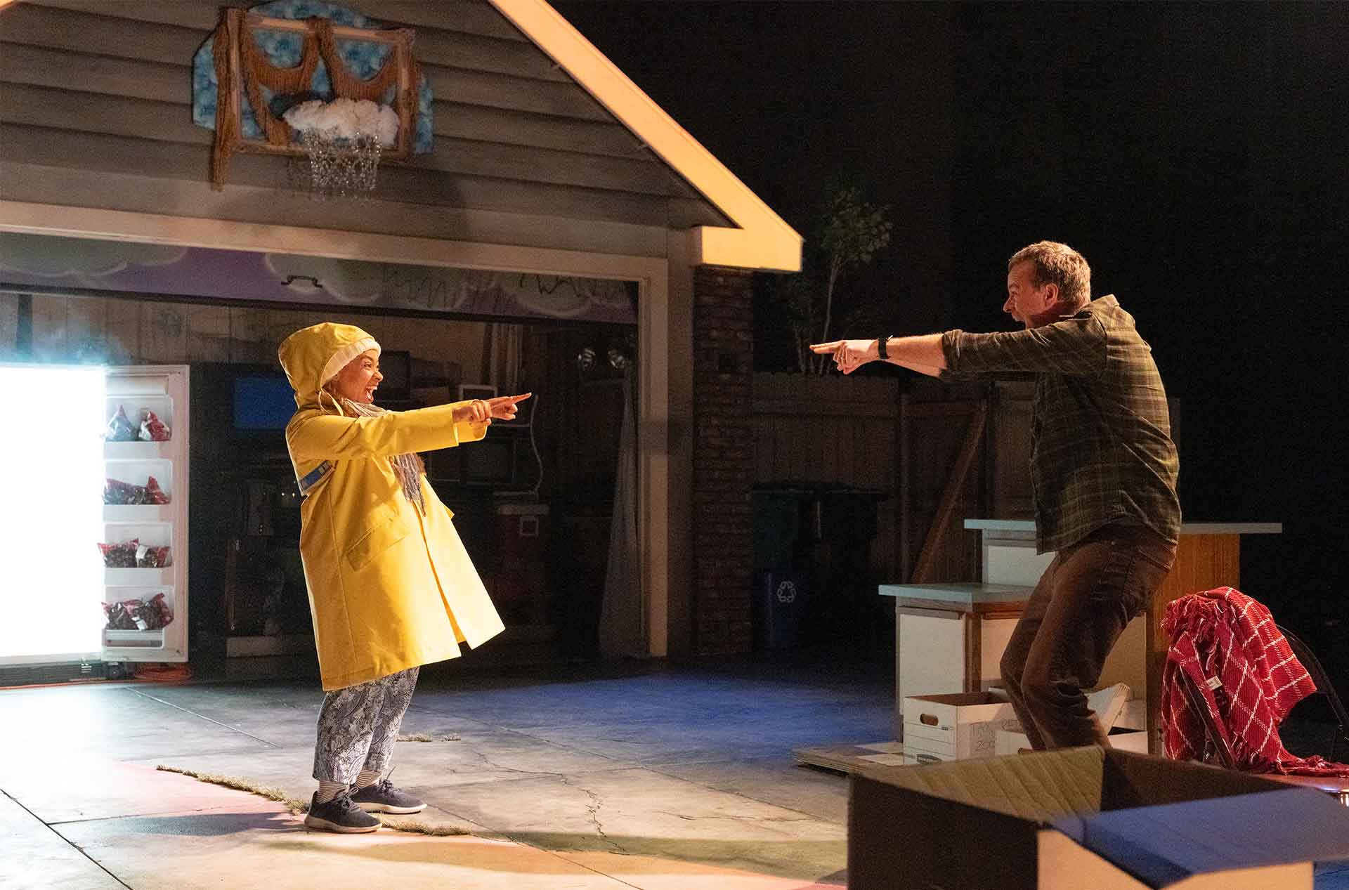 Two people face each other in a driveway set pointing playfully with matching gestures. An open refrigerator glows in the background, adding a surreal element to the scene.