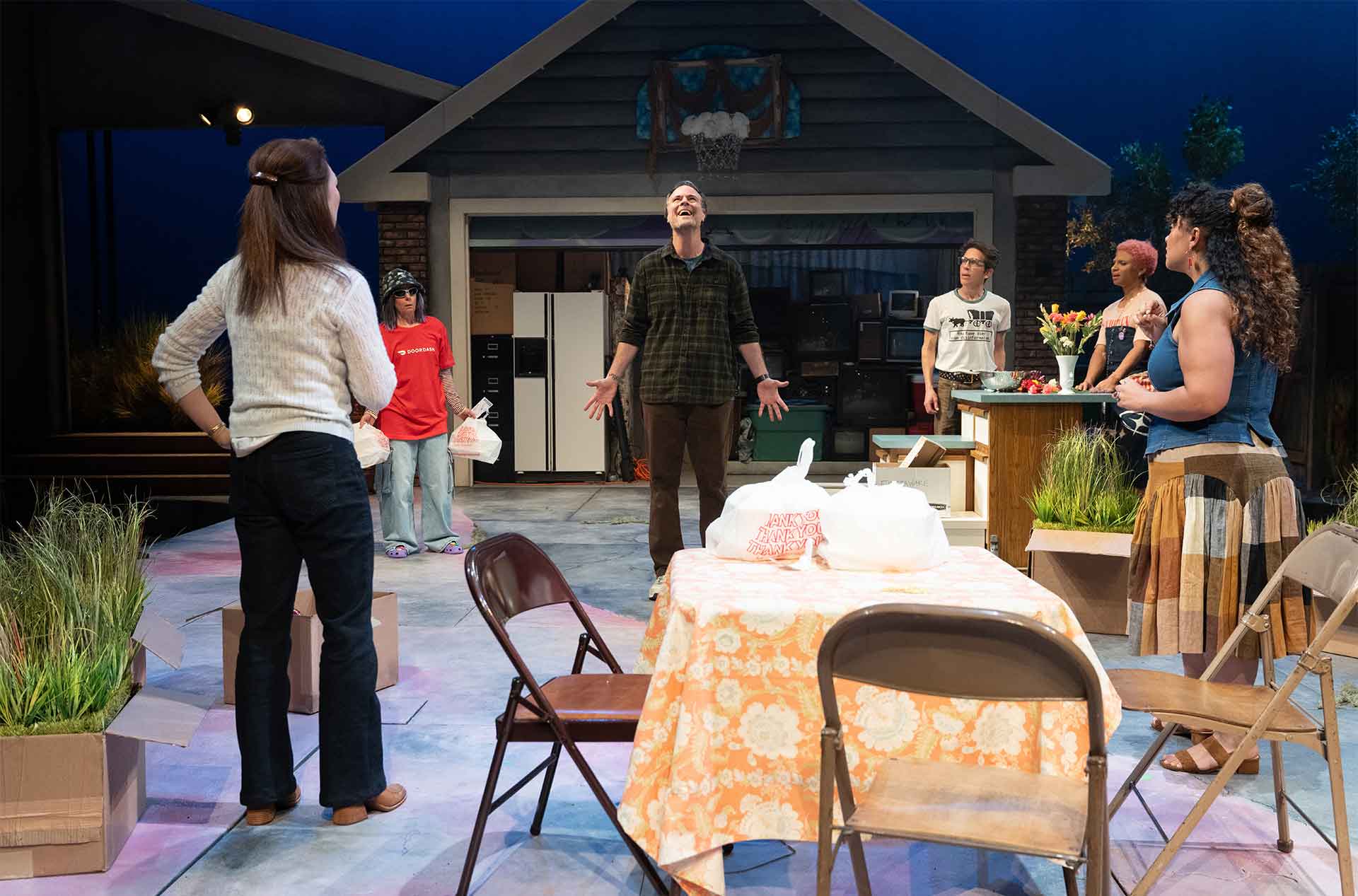 Group of people gather around a table in a driveway set as one person stands with arms open looking upward. Folding chairs, takeout bags, and household items suggest a casual gathering turned dramatic moment.