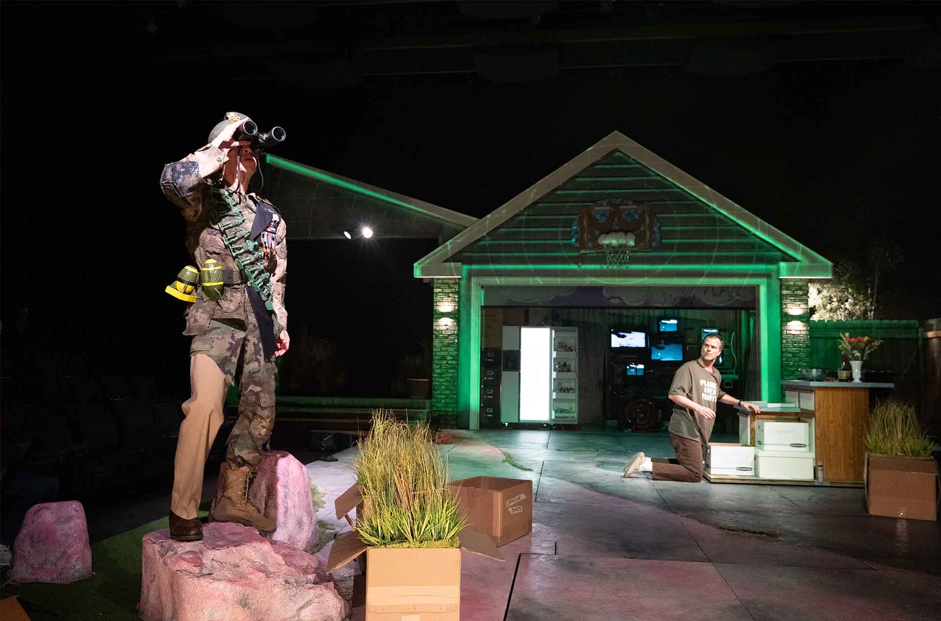 Person in a military style outfit stands on a rock looking through binoculars while another kneels nearby in a driveway set. Green lighting washes over the garage and scattered boxes around them.