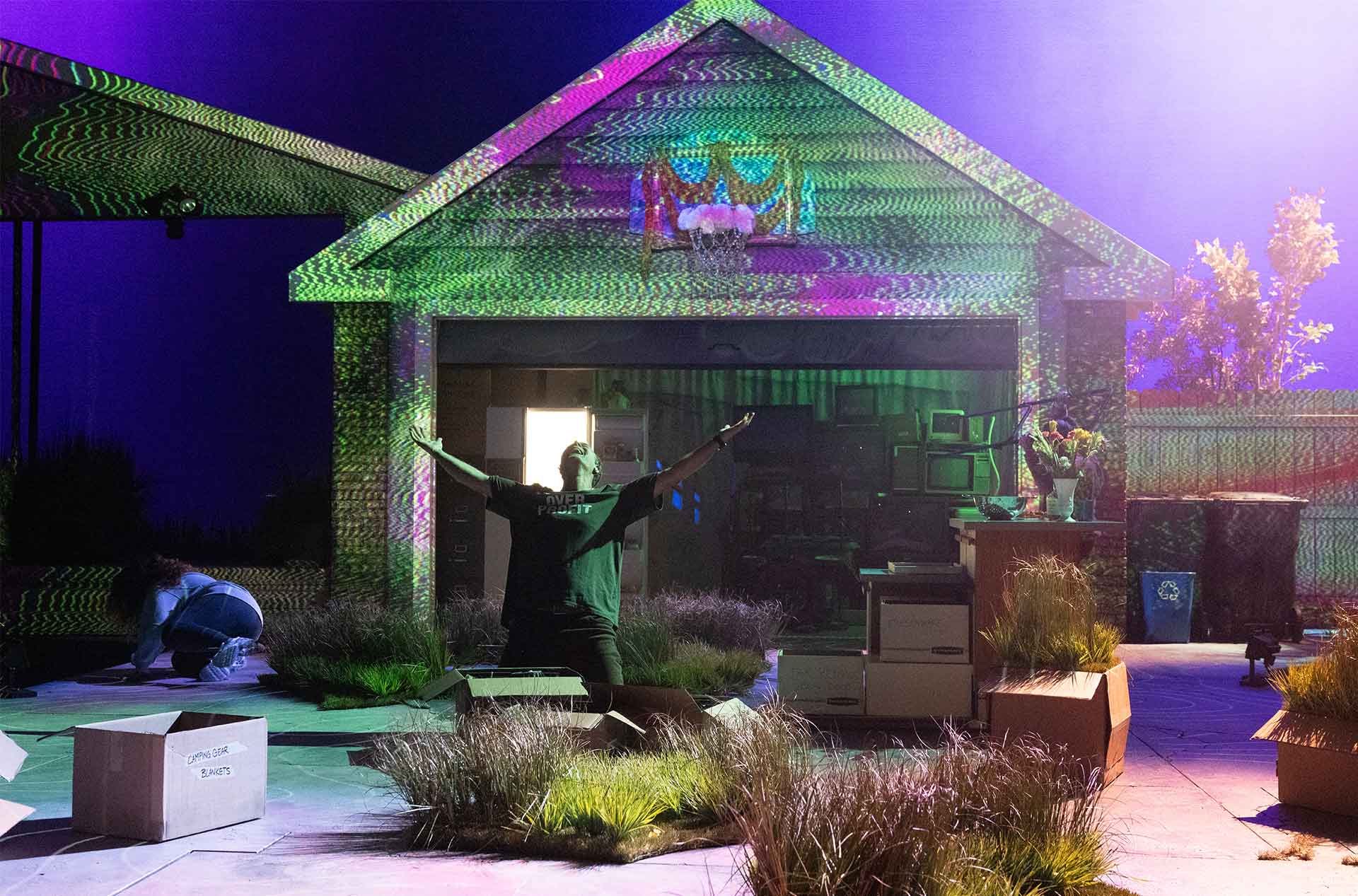 Person kneels with arms raised under shifting colorful light patterns projected onto a house facade. Cardboard boxes and plants surround the driveway stage creating a surreal atmosphere.