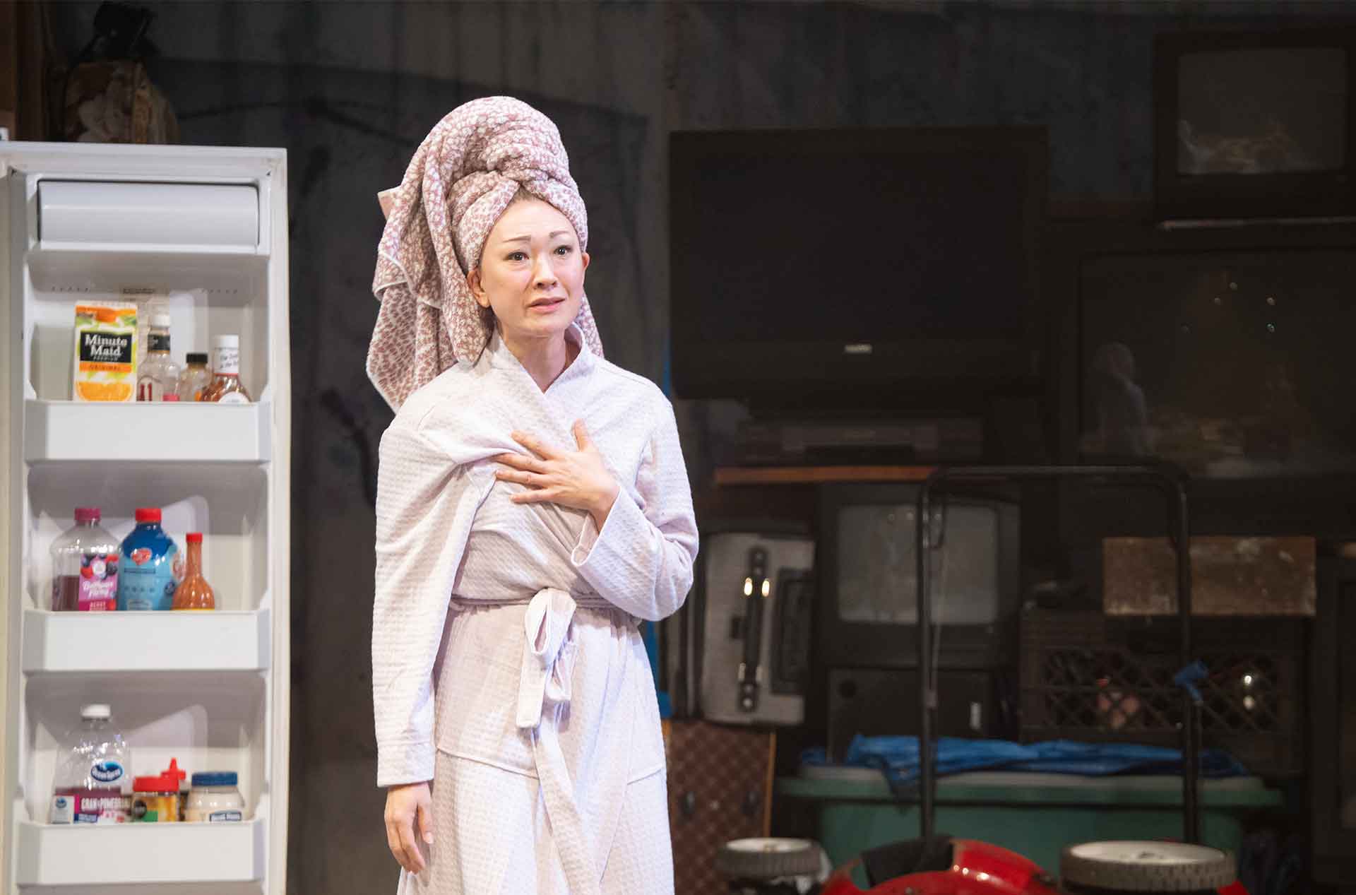 Person in a bathrobe and towel wrapped hair stands near an open refrigerator inside a garage set. Bottles and containers are visible in the fridge while the background shows household clutter.