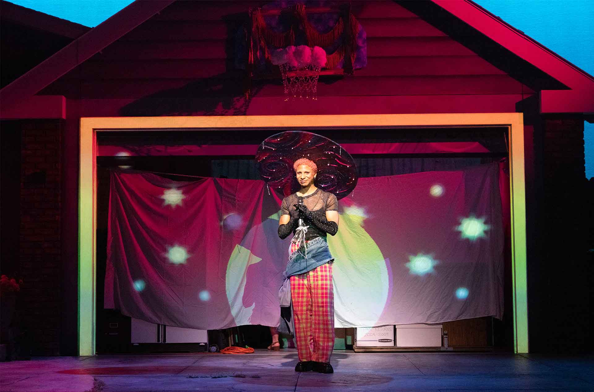 Performer stands centered in a driveway stage wearing bold layered clothing and a large sculptural hat under colorful lighting. A fabric backdrop with projected shapes creates a theatrical mood behind them.
