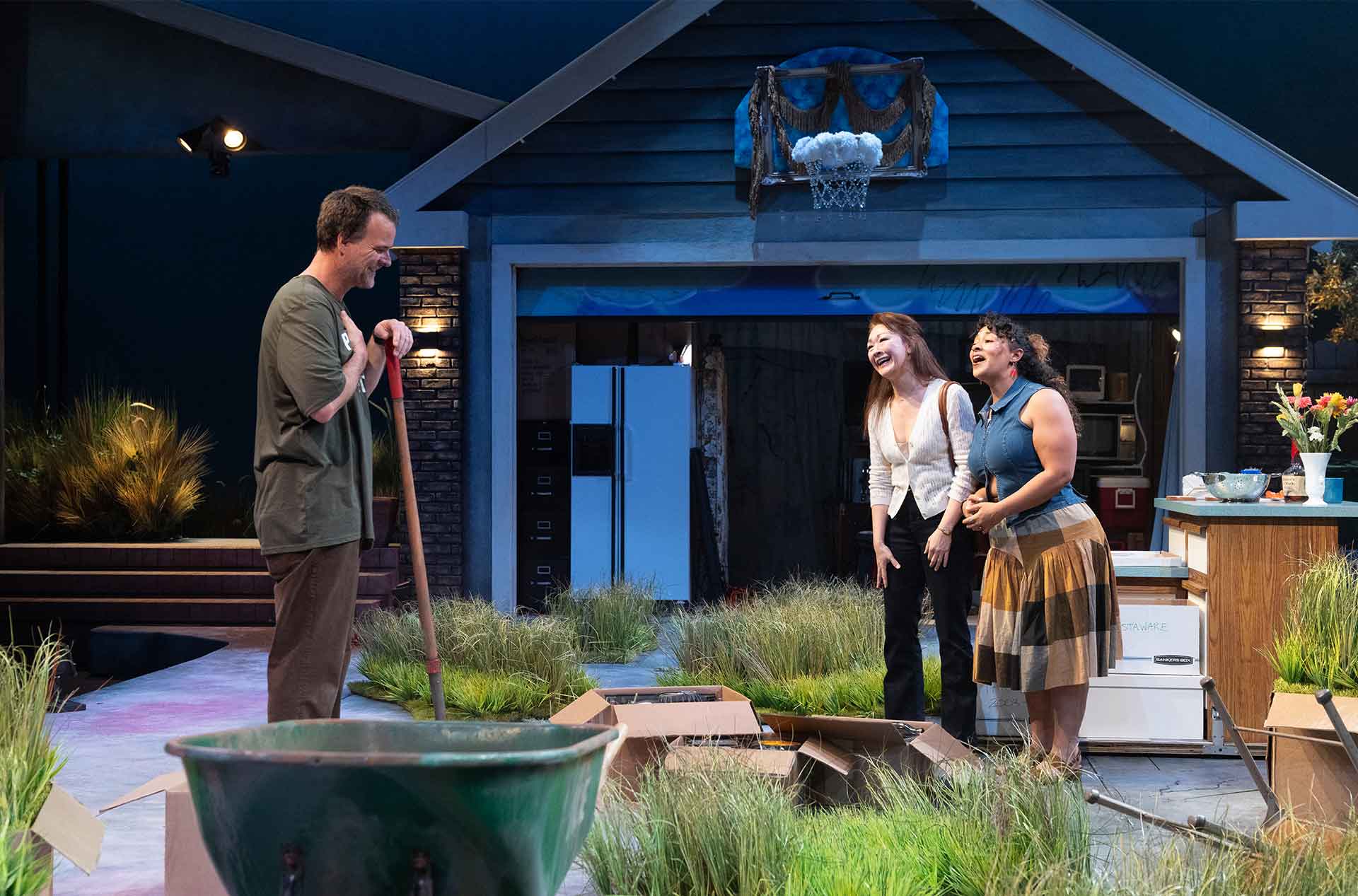 Three people stand in a driveway set smiling and leaning toward each other near scattered boxes and a wheelbarrow. The mood feels light and conversational in a staged suburban environment.