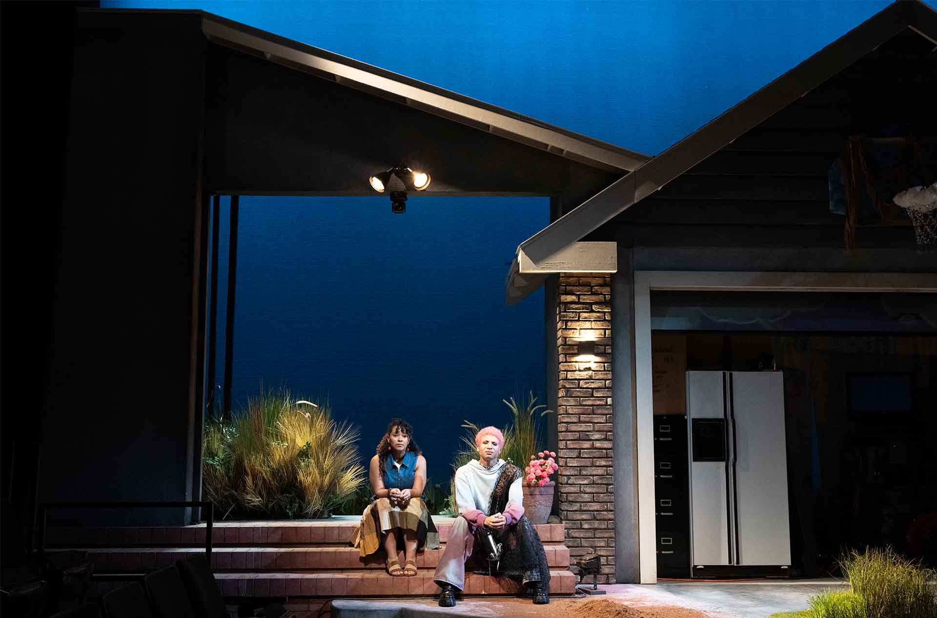 Two people sit side by side on front steps of a house set at night with stage lighting and tall grass around them. The scene feels quiet and reflective in a suburban driveway setting.