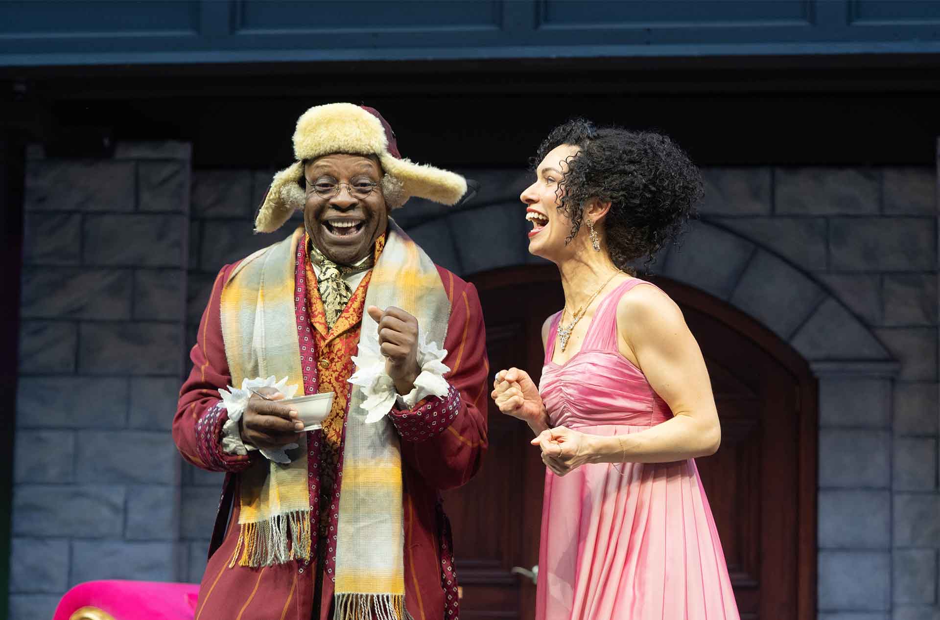 Two performers laugh together on a theater stage. One wears a fur lined hat and red coat while holding a teacup and the other wears a pink dress.