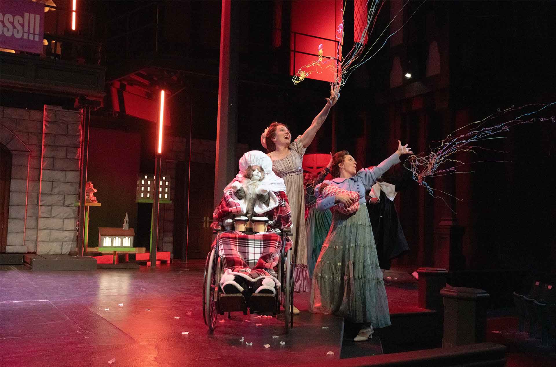 Performers toss colorful streamers across a dark theater stage lit in red. One performer sits in a wheelchair covered with a plaid blanket while others reach upward beside them.