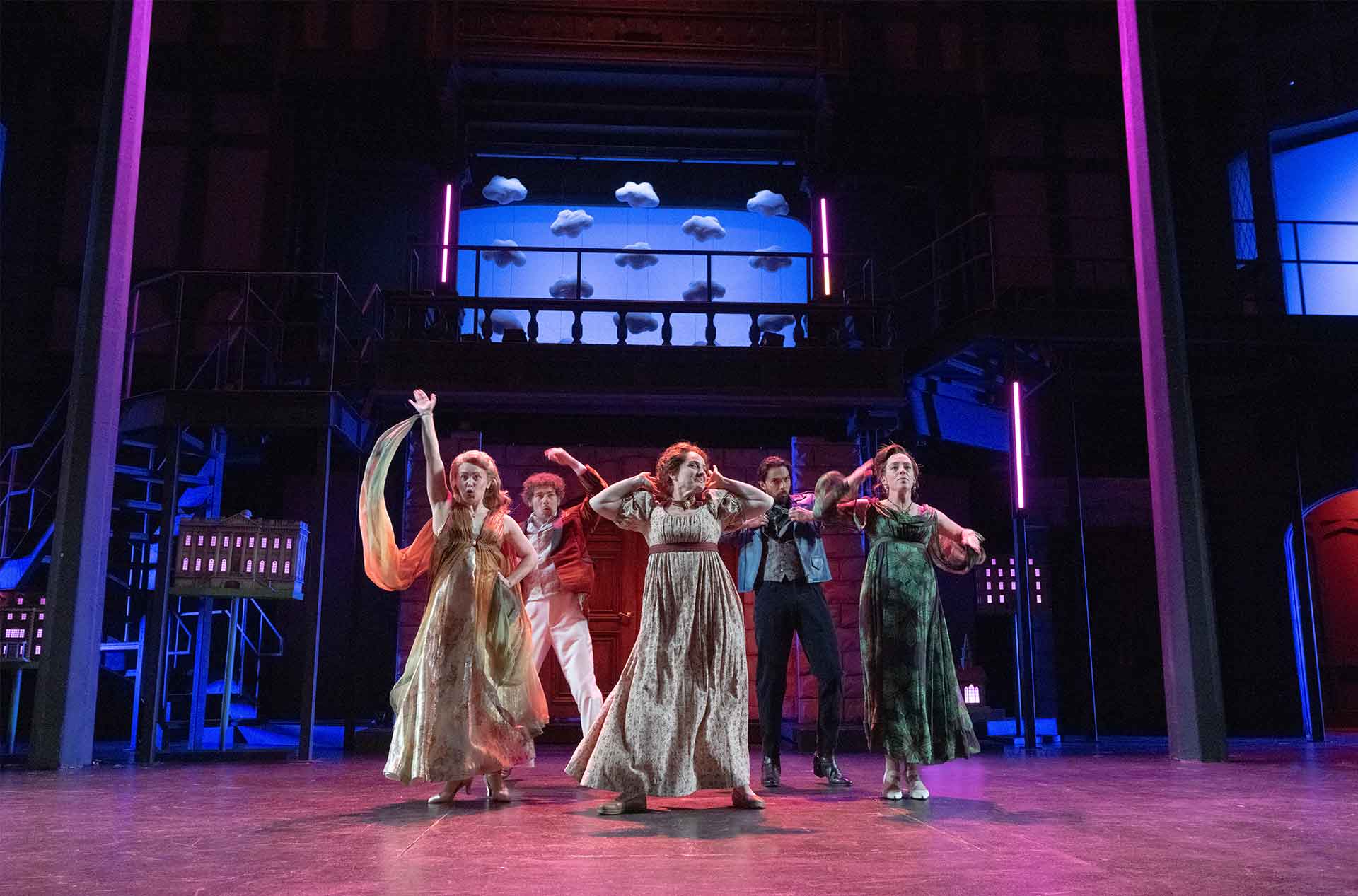 Five performers dance on a theater stage beneath a balcony lined with cloud props. Blue and pink stage lights frame the scene.