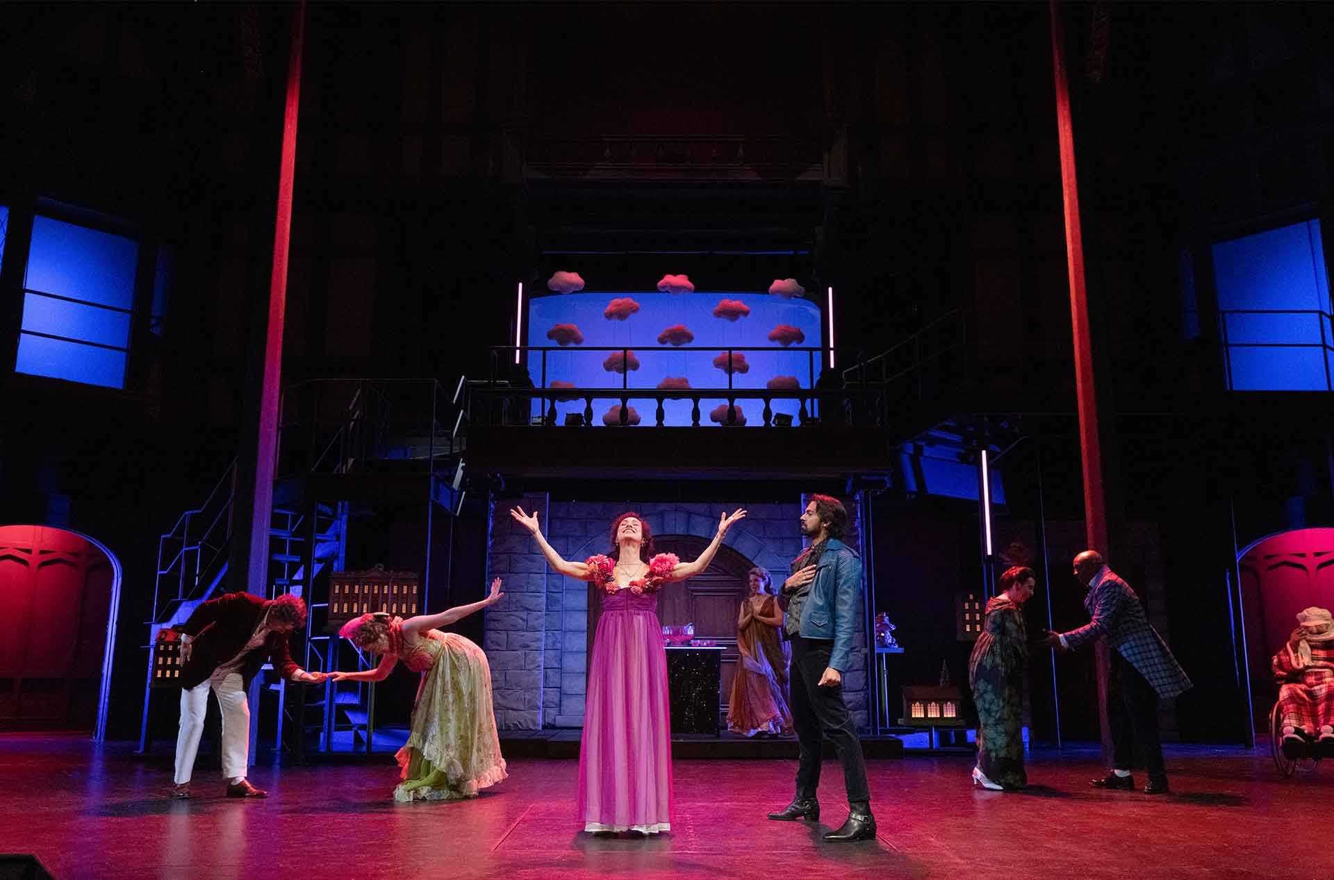 A theater ensemble performs under dramatic red and blue lighting. A performer in a pink dress stands center stage with arms raised while others pose around them.