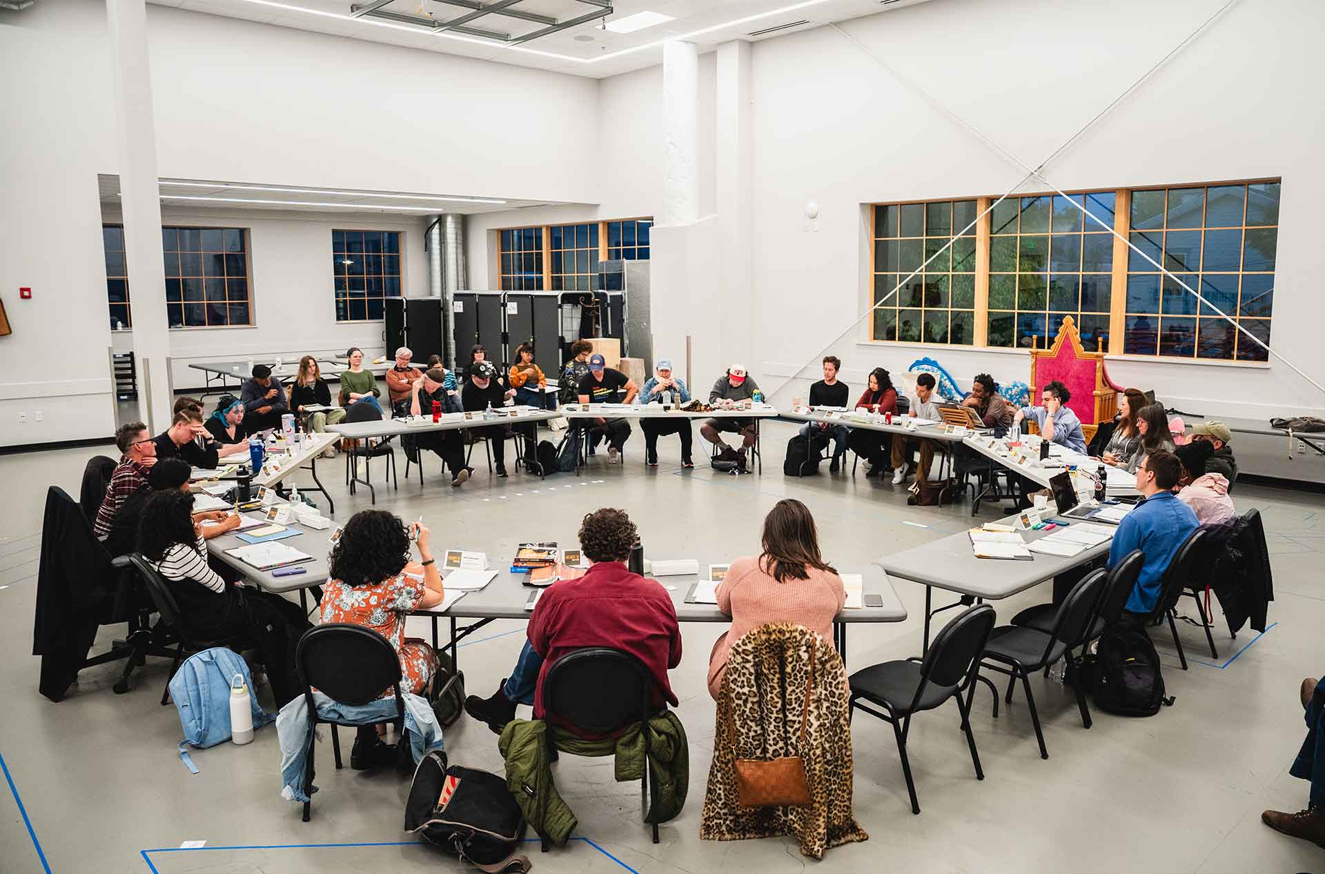 Wide view of a cast seated in a circle during a play rehearsal inside a bright rehearsal hall.