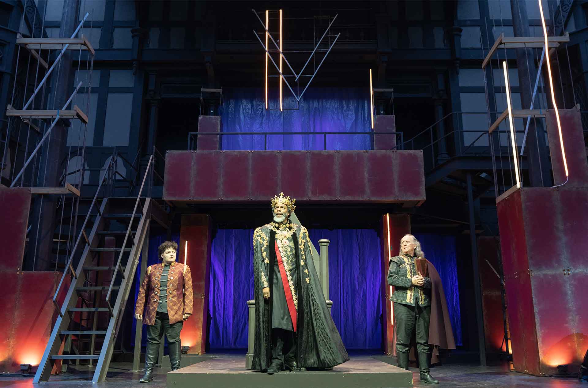 Three performers stand on a raised platform beneath blue and purple stage lighting. The center performer wears a gold crown and ornate black robe in front of a throne.