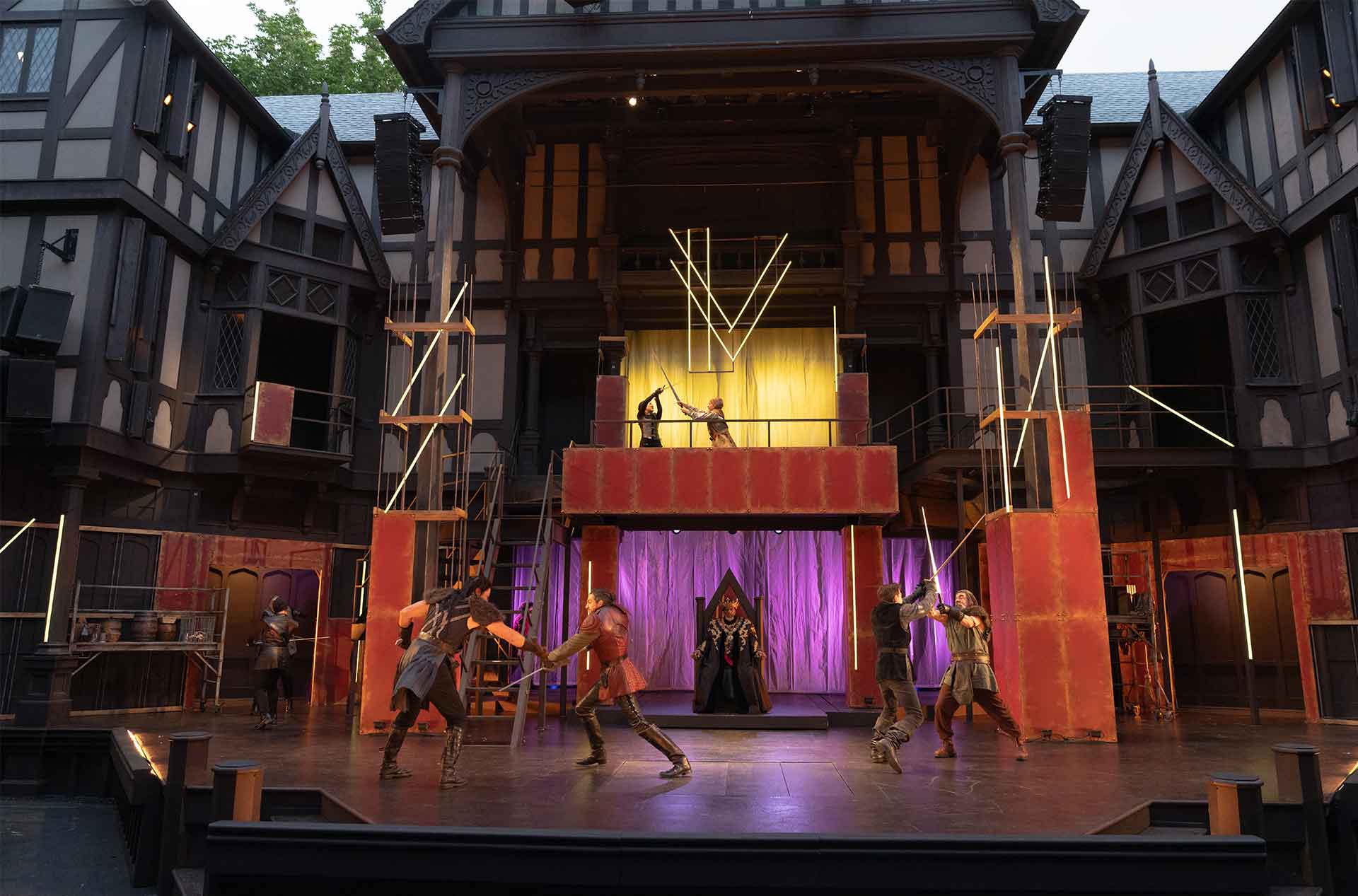 Performers duel with swords across a multi level outdoor stage. The set has Tudor style architecture.