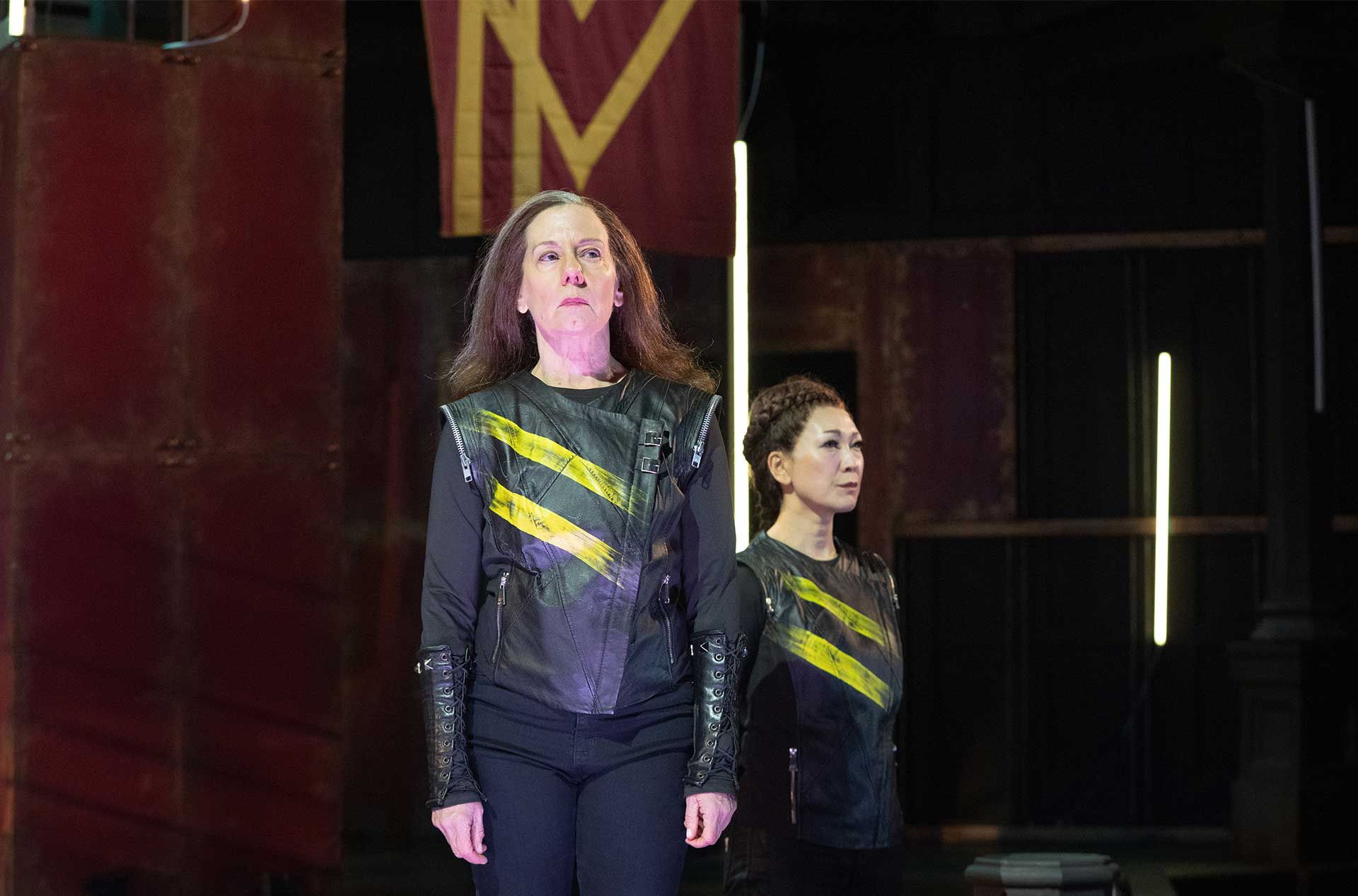 Two performers stand in matching black costumes with yellow diagonal stripes. Red set pieces and vertical light bars create a stark stage backdrop.