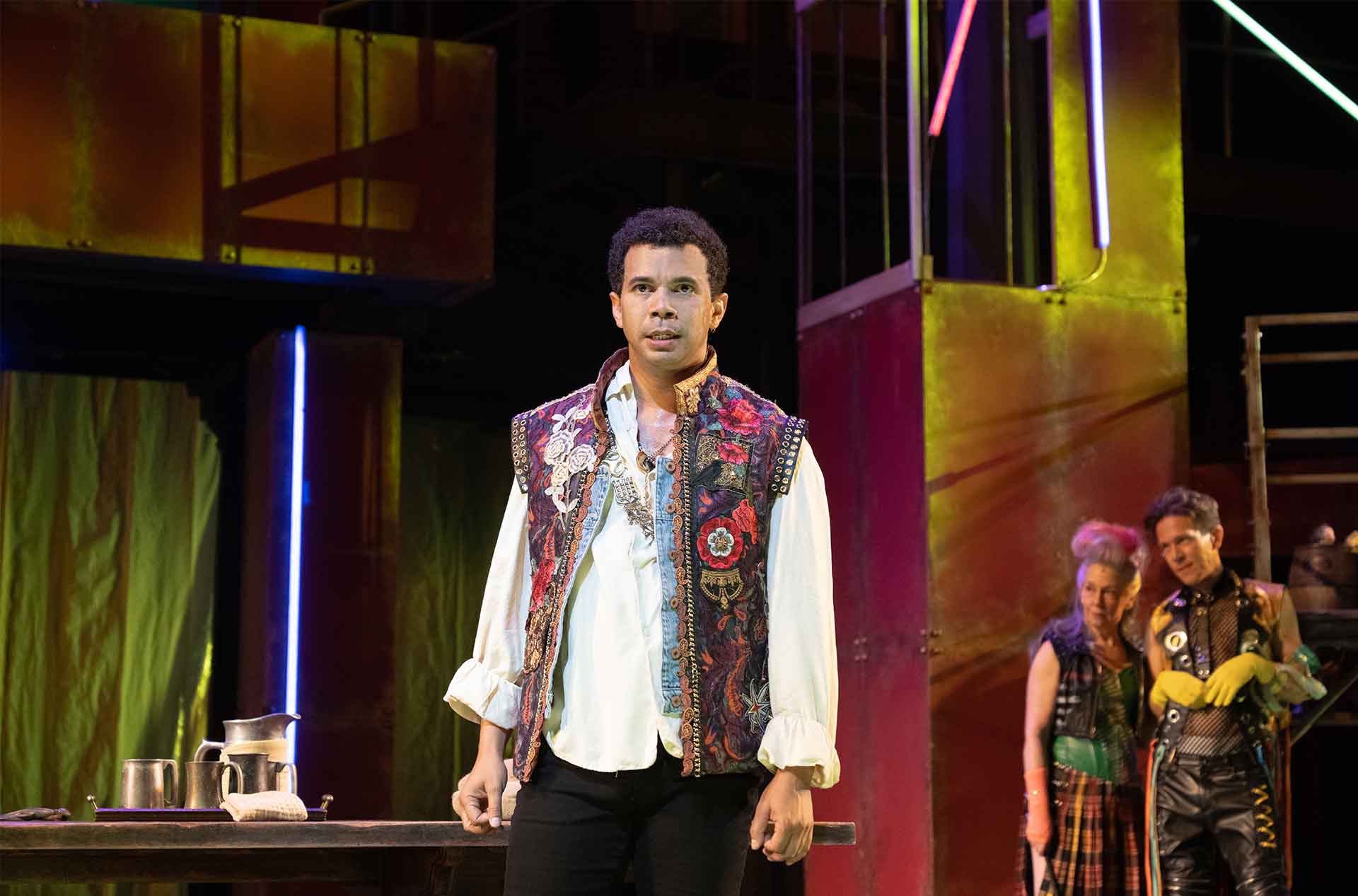 A performer in a floral vest stands near a table with metal pitchers while two others watch from the background. The stage is lit with saturated green and red tones.