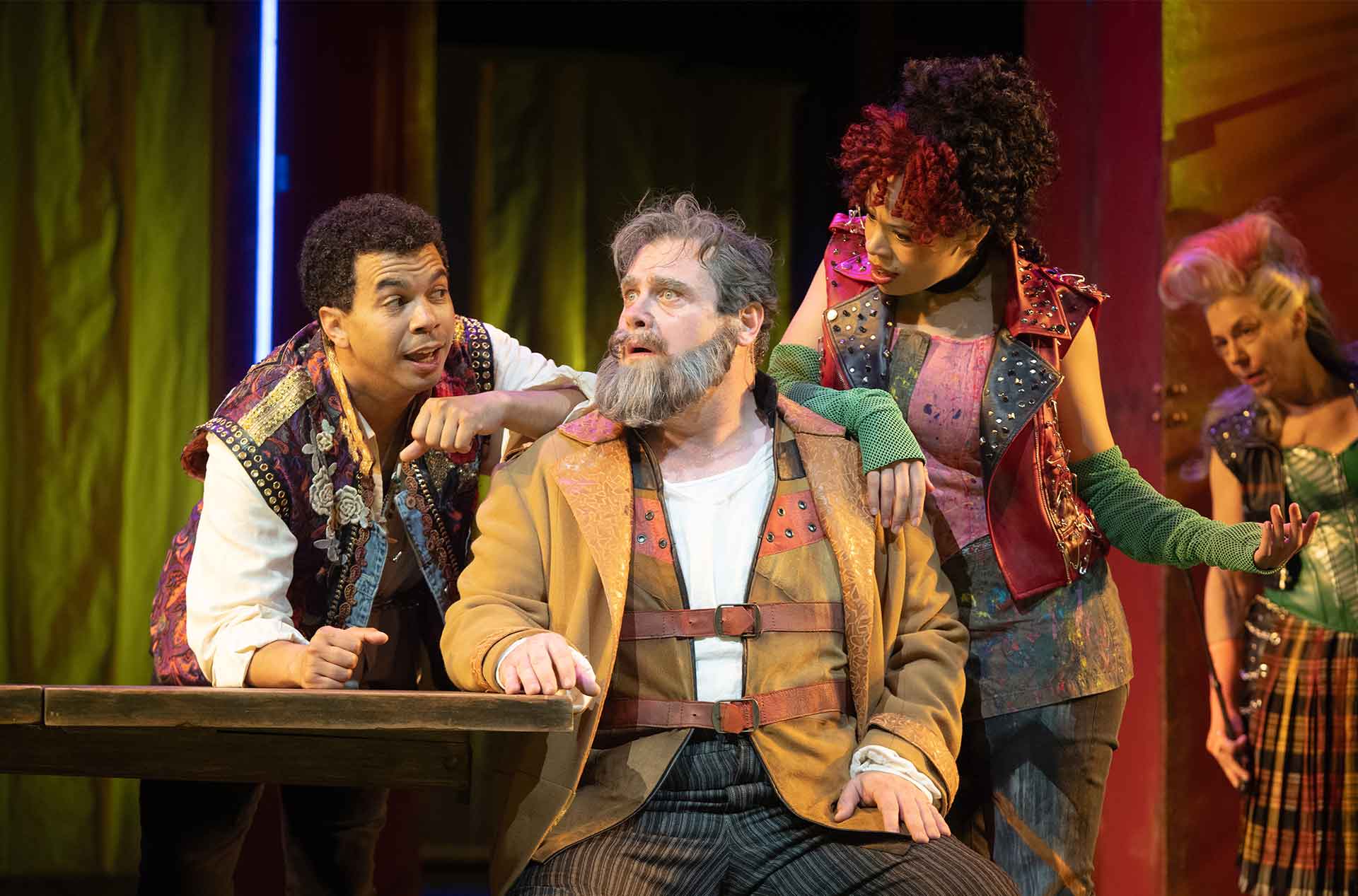 Three performers gather closely around a wooden table during a tense stage scene. Bright green and red lighting highlights the layered costumes and expressive reactions.