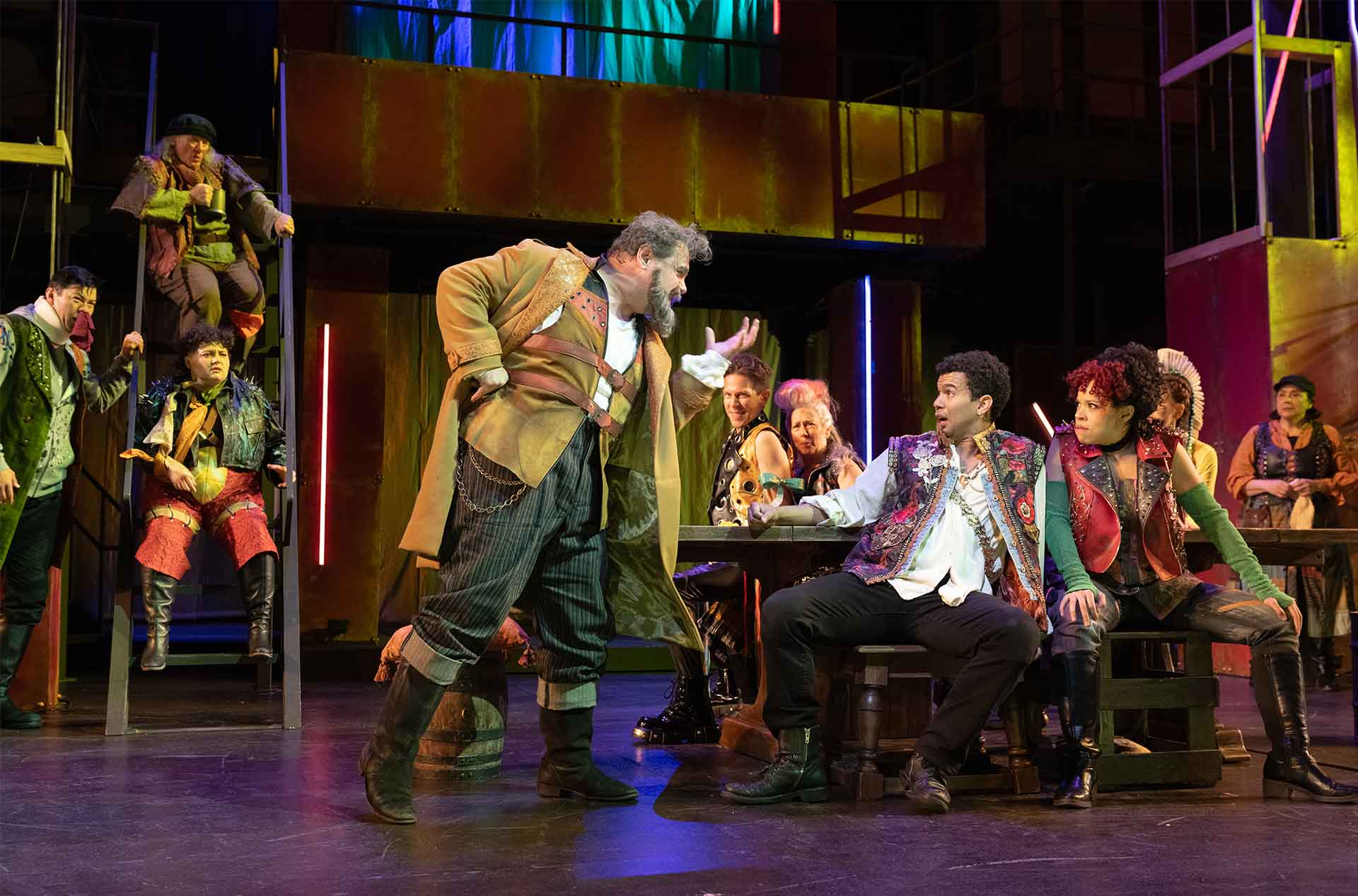 A large ensemble fills the stage in colorful theatrical costumes. One performer gestures toward another seated at a wooden table while others watch from ladders and benches.