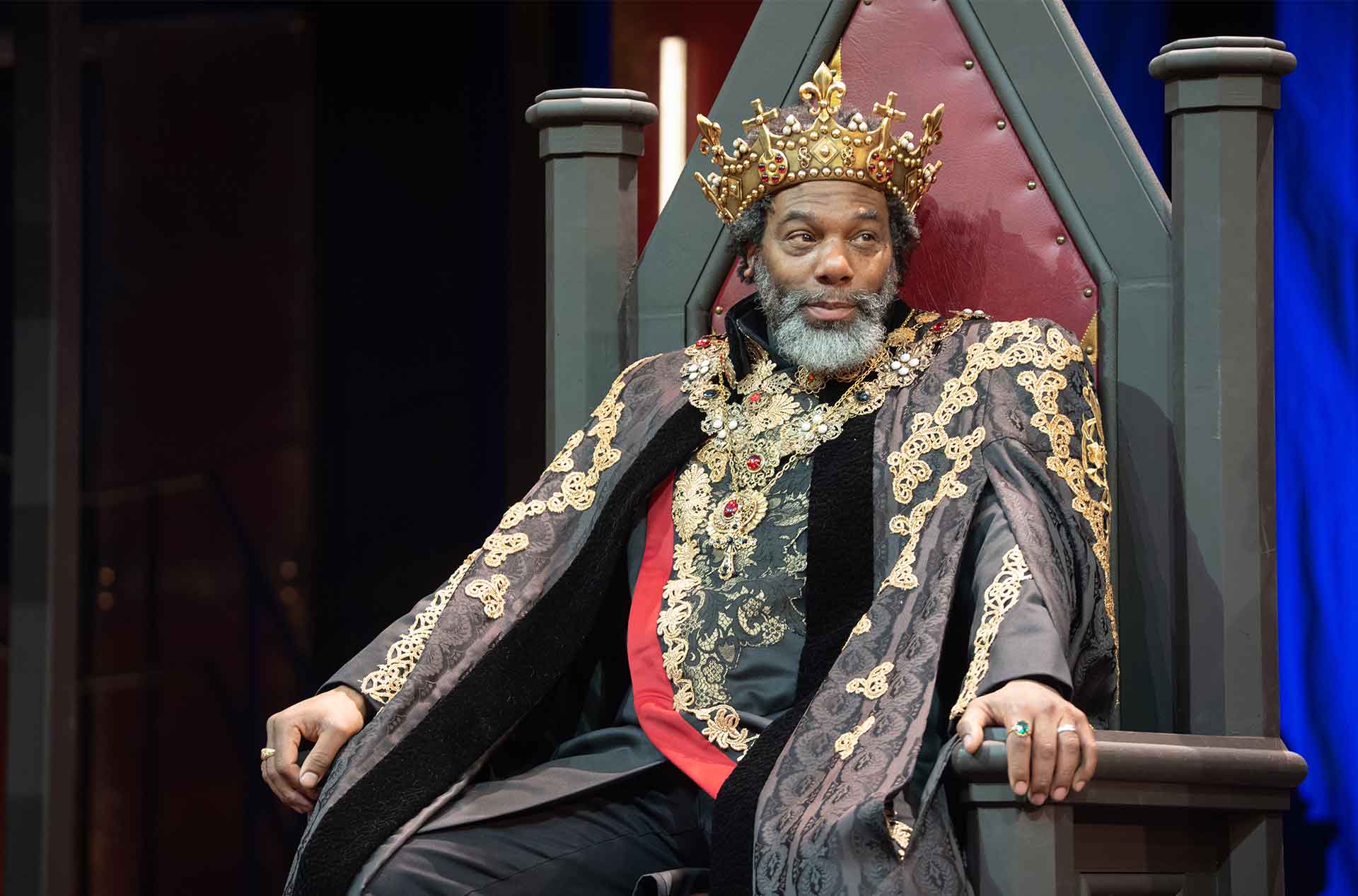A crowned performer sits on a tall throne wearing an ornate black robe with gold embroidery and jeweled details. 