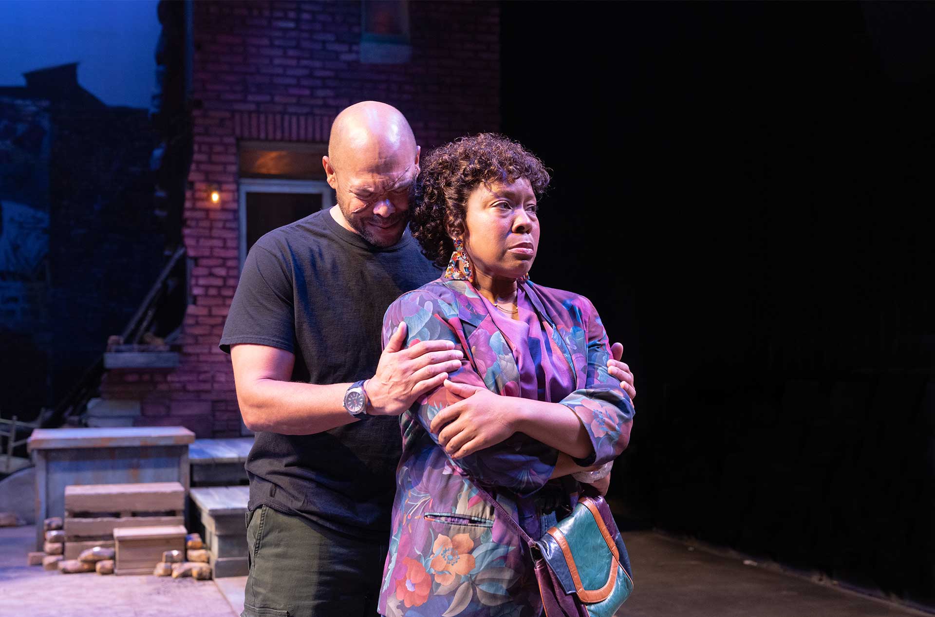 One performer stands behind another and gently holds their shoulders. Both appear distressed against a brick courtyard set.